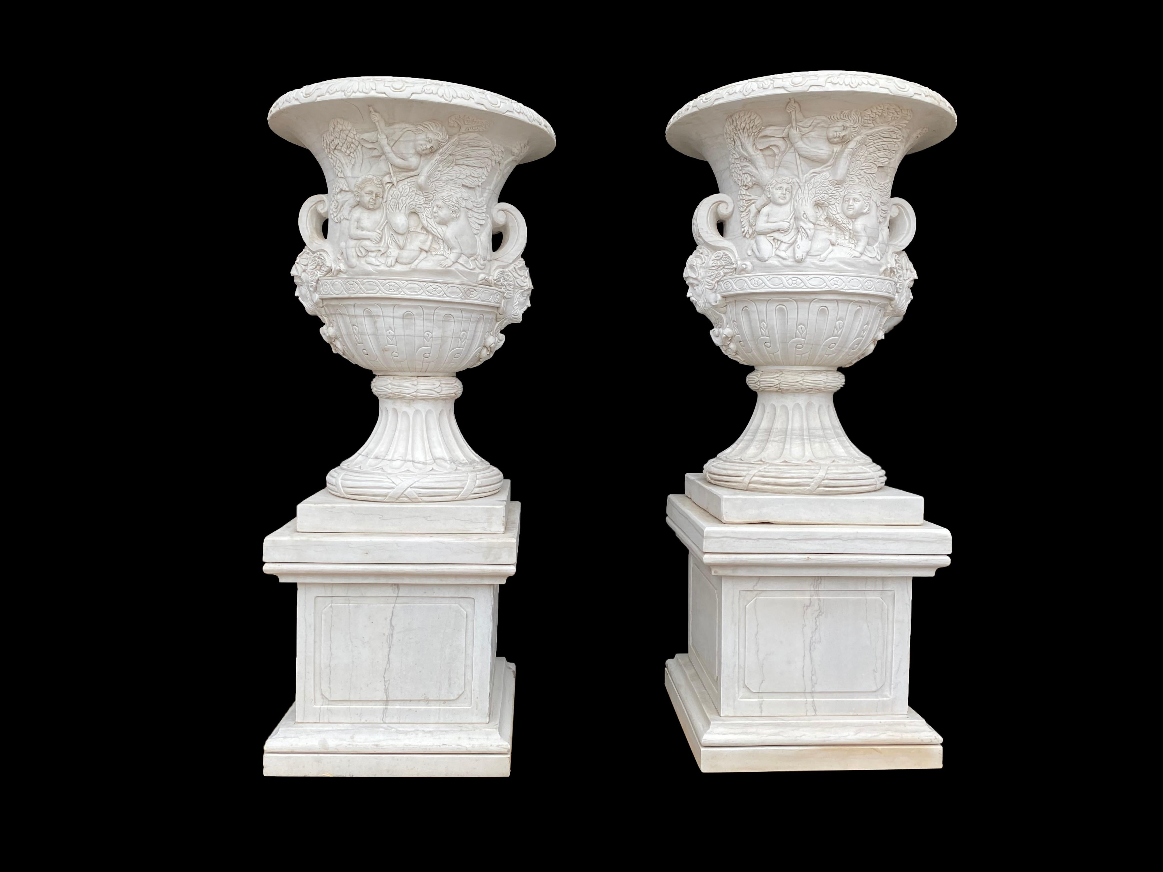 A beautiful pair of 21st century white marble large campana-form urns with figural friezes. The body with carved foliate scrolled decoration and mask handles, raised on square plinth bases, mounted on square pedestals. The pair have stunning carved