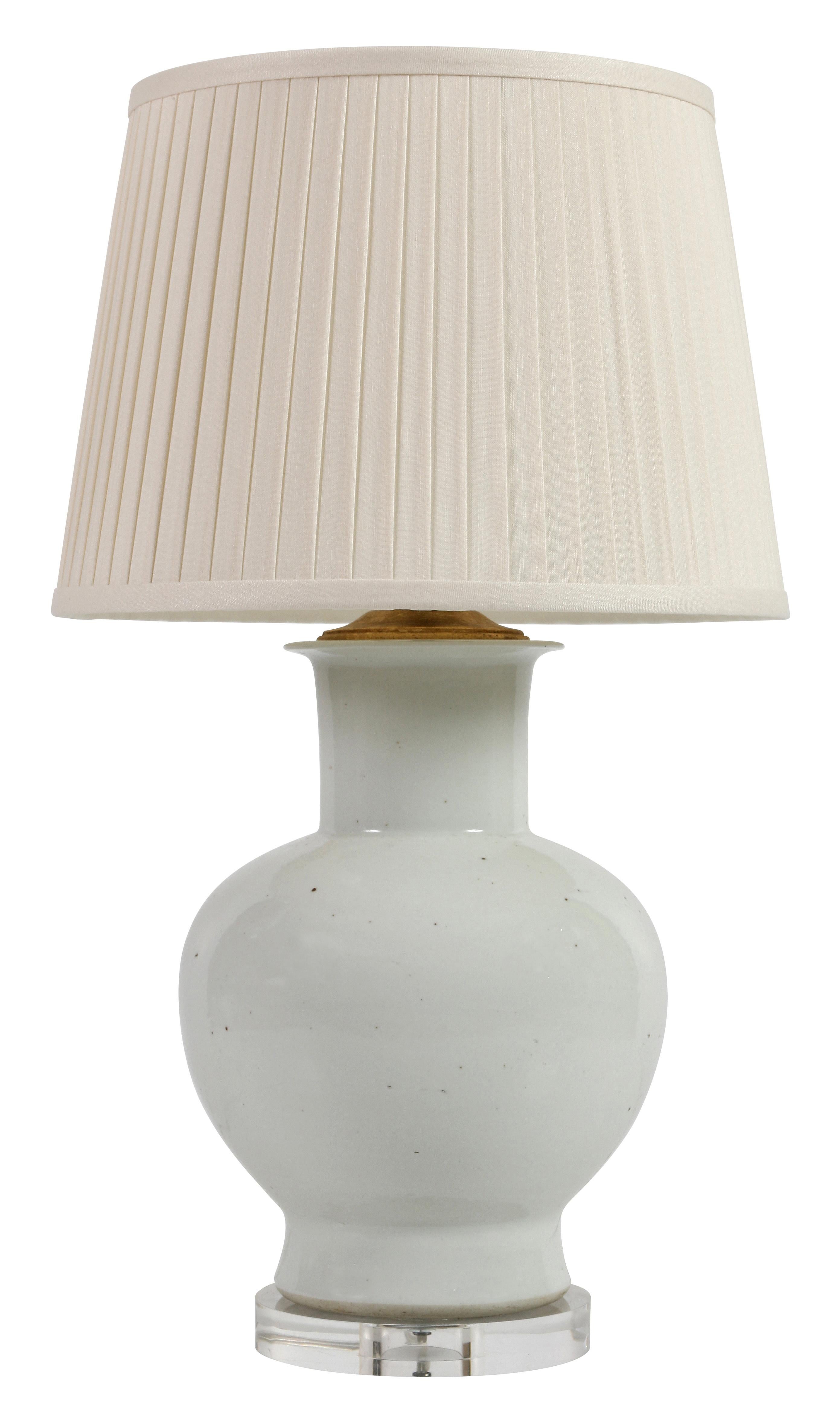 A pair of white ceramic urn-shaped lamps with brass fittings on a lucite base with a pleated linen drum shade. With their classic shape and neutral color, these lamps can go anywhere and can add a bit of freshness to a room.