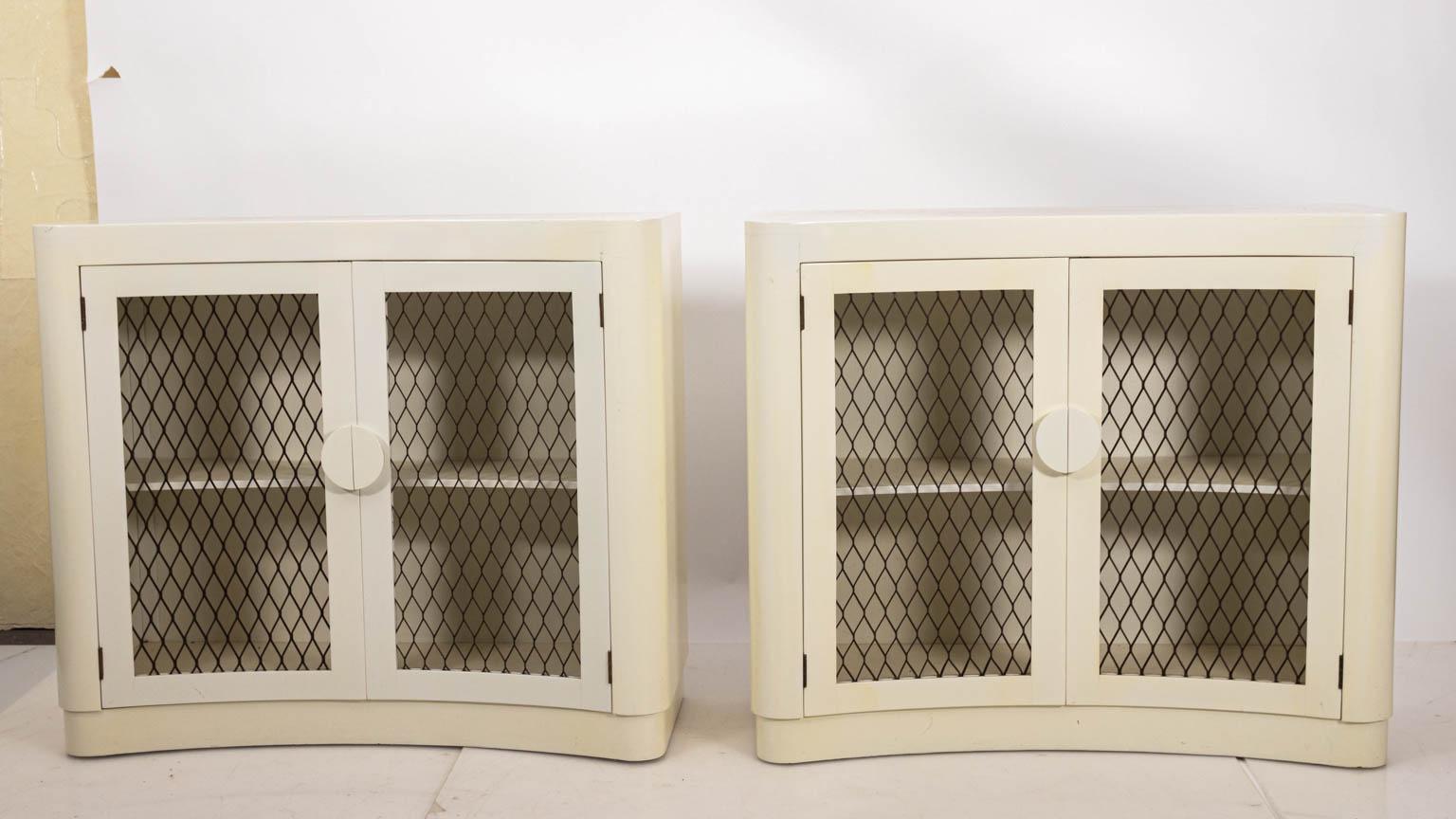 Pair of white lacquered Hollywood Regency style cabinets with metal mesh insert doors, circa 1940s. One shelf is featured on the inside of each piece. Please note of wear consistent with age including cracking and crazing to the finish. Made in the