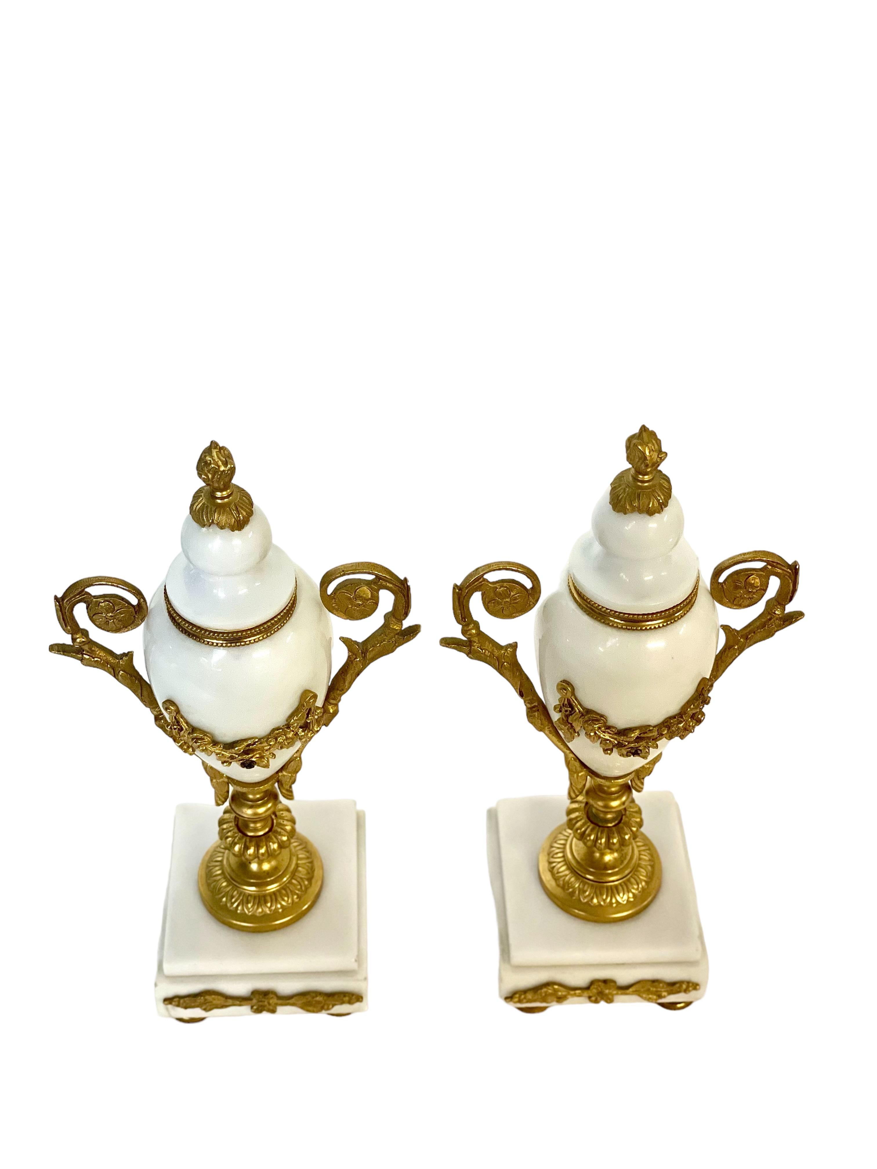 A pair of highly ornate 'Cassolettes' (decorative urns) crafted from white marble mounted with ormolu (finely gilded bronze) and designed to sit atop a mantlepiece, perhaps on either side of a matching clock. Designed to resemble a classically