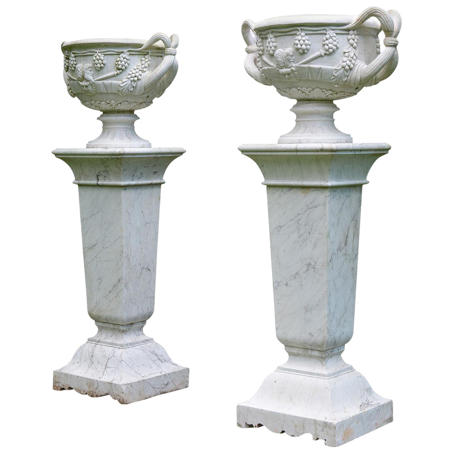 A pair of carved marble urns, each with double-looped handles, the urn body with portrait busts and grapevines, the motifs and overall shape of the urns inspired by the Warwick Vase, an ancient Roman urn discovered at Hadrian’s Villa, in Tivoli,