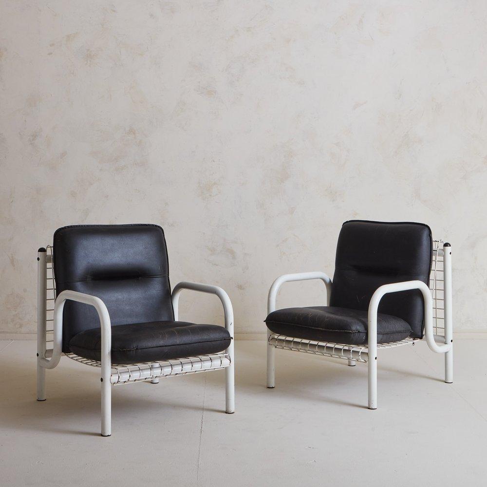 A pair of vintage French lounge chairs featuring tubular white metal frames with curved arms. These chairs have metal seats and backs in a perforated square pattern which elegantly drape over the frame. Each chair has two vegan leather cushions with