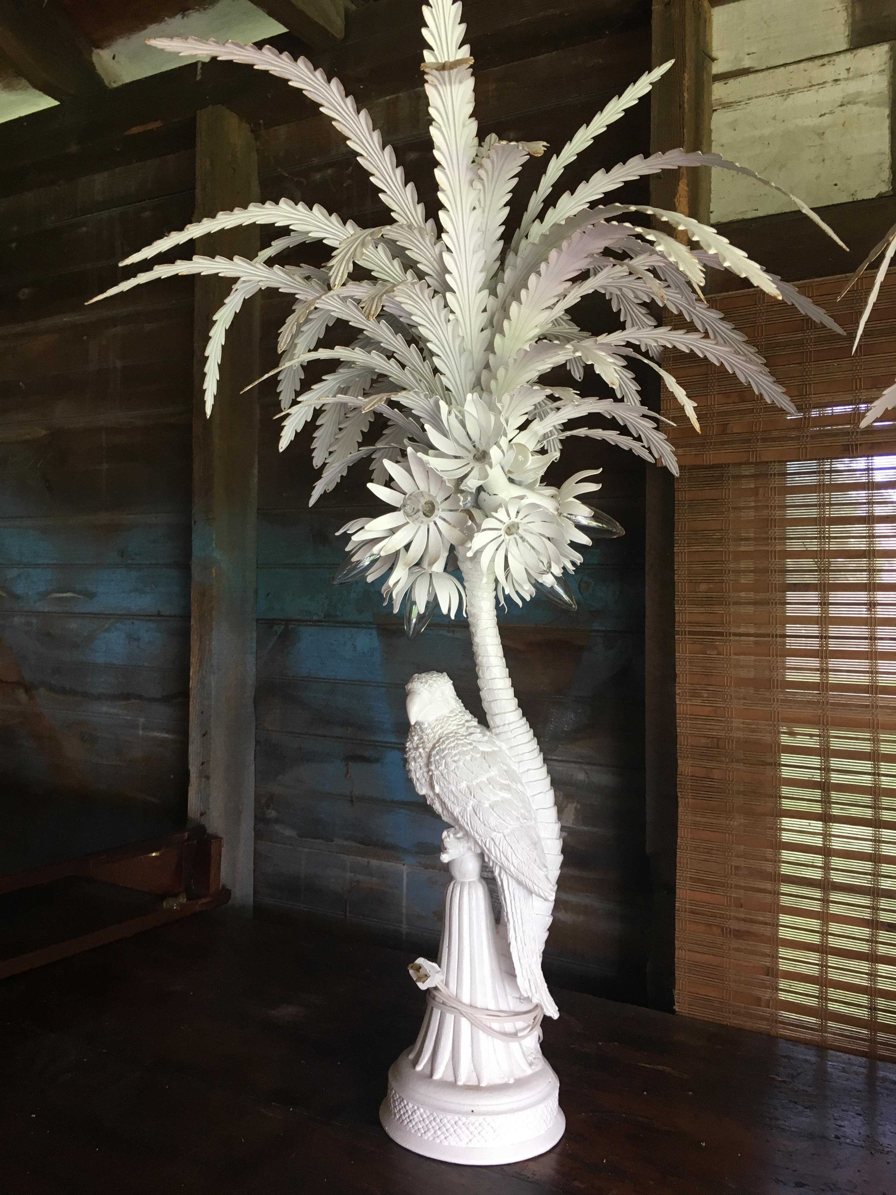 Pair of white painted tole and plaster parrot lamps with detachable glass drops. A parrot made in plaster sits atop a pedestal with a palm tree behind. Metal flowers extend out into metal palm branches with glass drops hanging off the ends. These