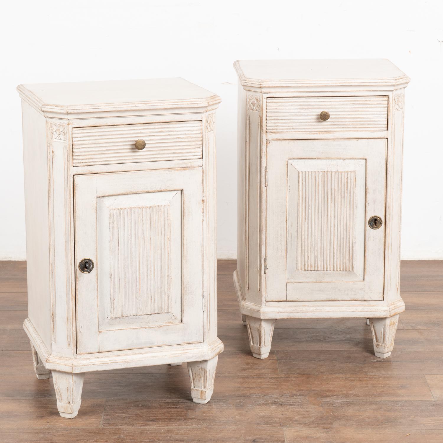 Pair, small Swedish country Gustavian style white painted nightstands or small cabinets standing on tapered feet.
Canted sides have simple fluted carving topped with floral medallion. Fluted carving accents the doors and drawers as well. 
Newer