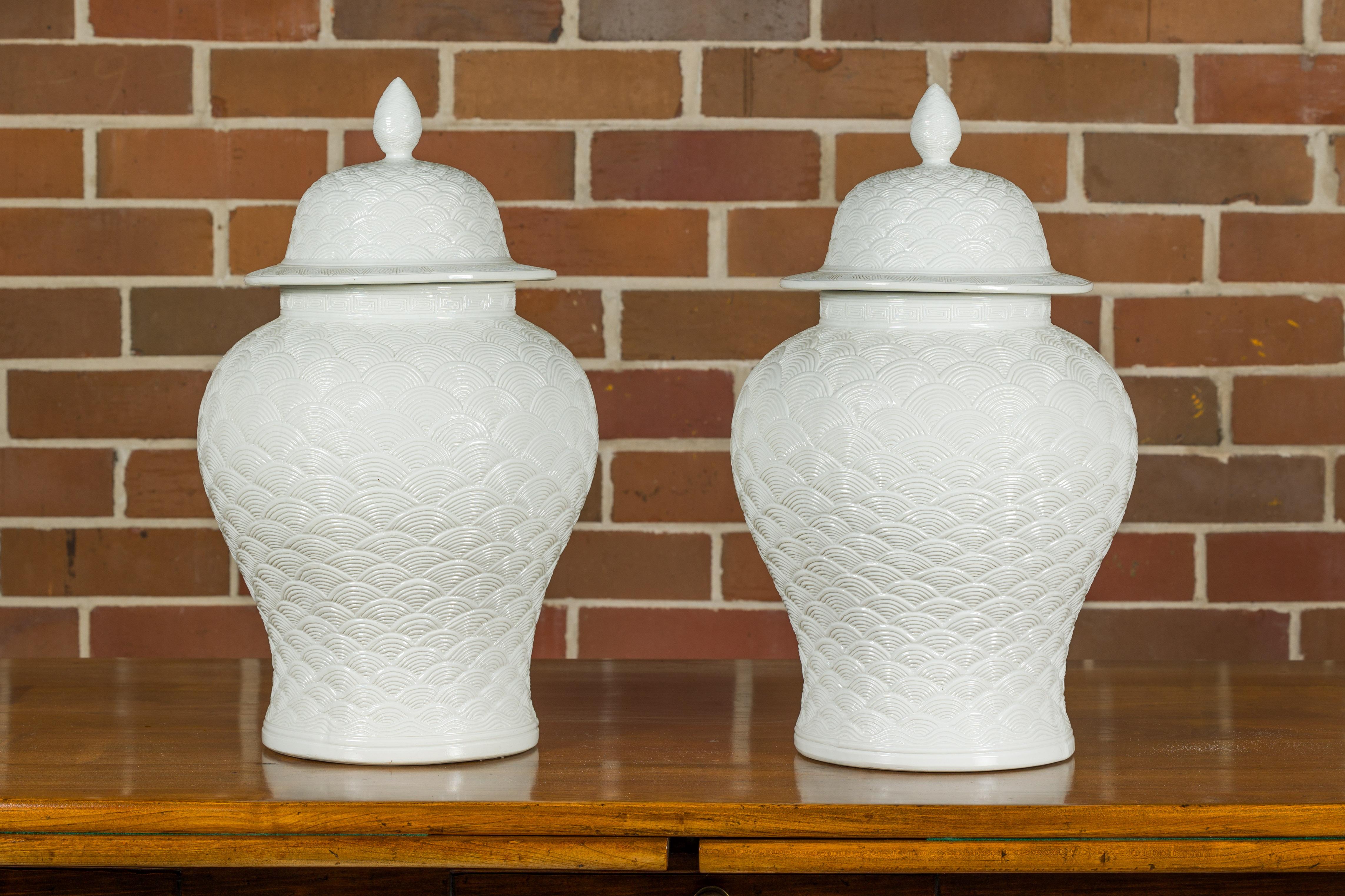 A pair of white porcelain fish scale lidded jars with finial at the top and Greek Key frieze on the neck. This pair of white porcelain lidded jars is a testament to refined elegance and intricate design. The body of each jar is adorned with a fish