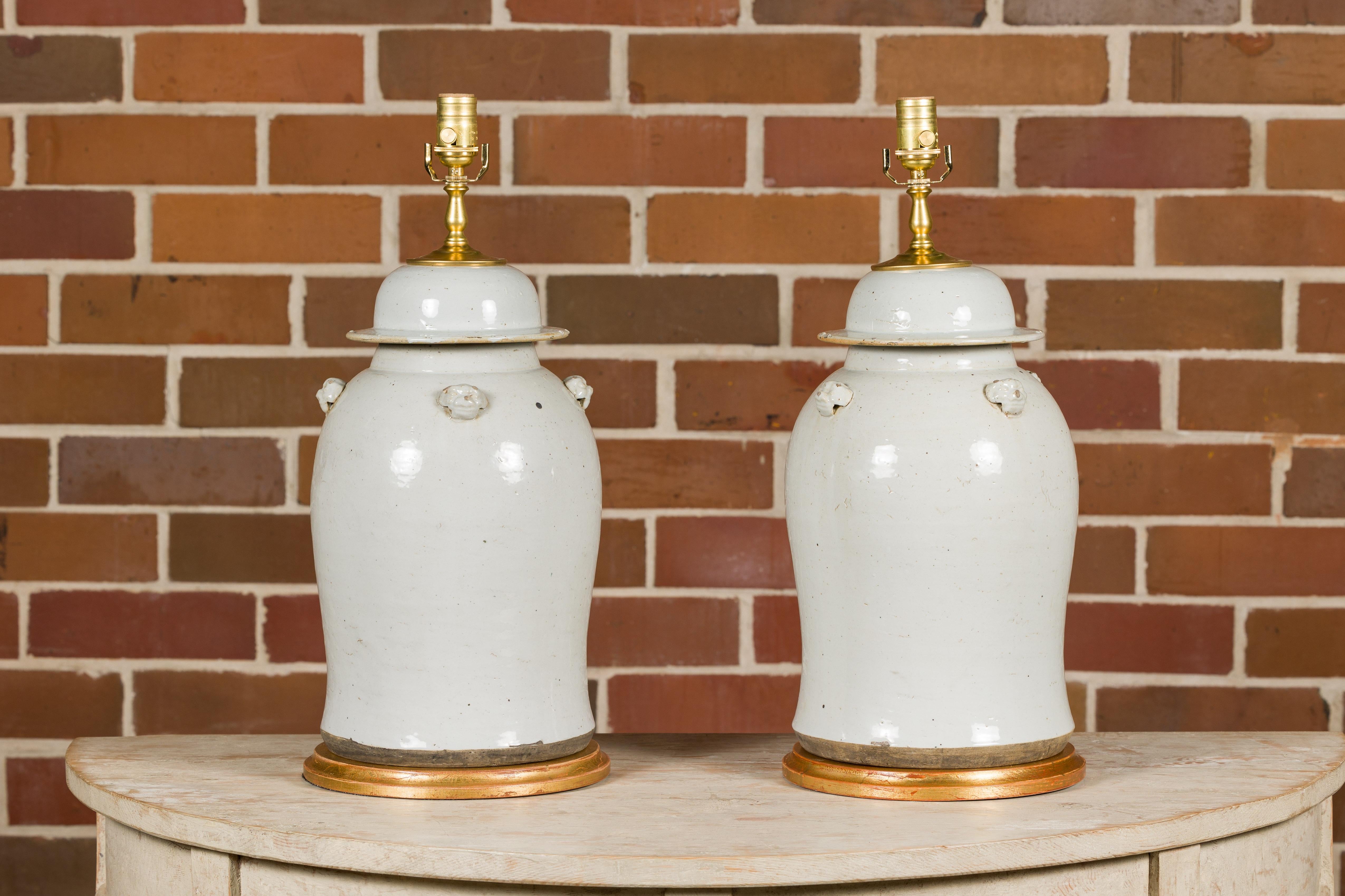 A pair of white porcelain lidded urns made into table lamps with single lights on circular gilded bases. Immerse your home in the elegant simplicity and timeless beauty of this exquisite pair of white porcelain lidded urns transformed into table