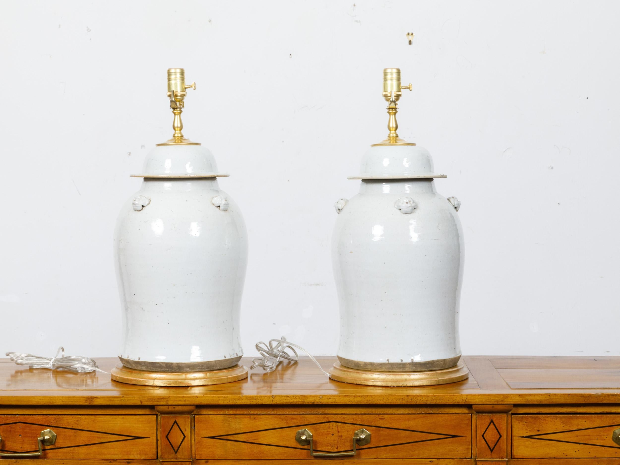 A pair of white porcelain lidded urns made into table lamps with single lights on circular gilded bases. This elegant pair of table lamps, crafted from white porcelain urns, epitomizes timeless sophistication. The urns feature slender necks and