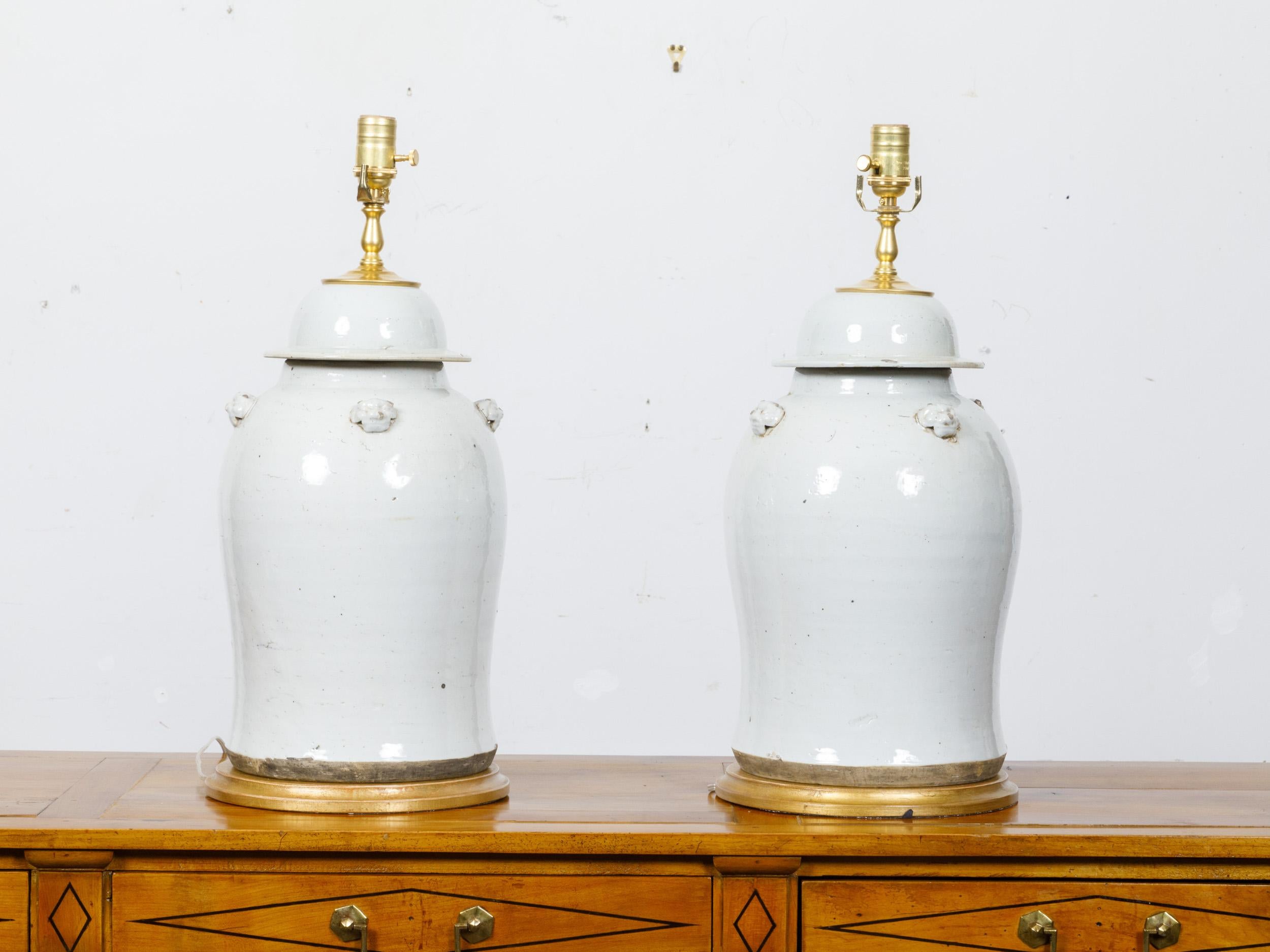 A pair of white porcelain lidded urns table lamps with single lights on circular gilded bases, wired for the USA. This elegant pair of table lamps, crafted from white porcelain urns, epitomizes timeless sophistication. The urns feature slender necks