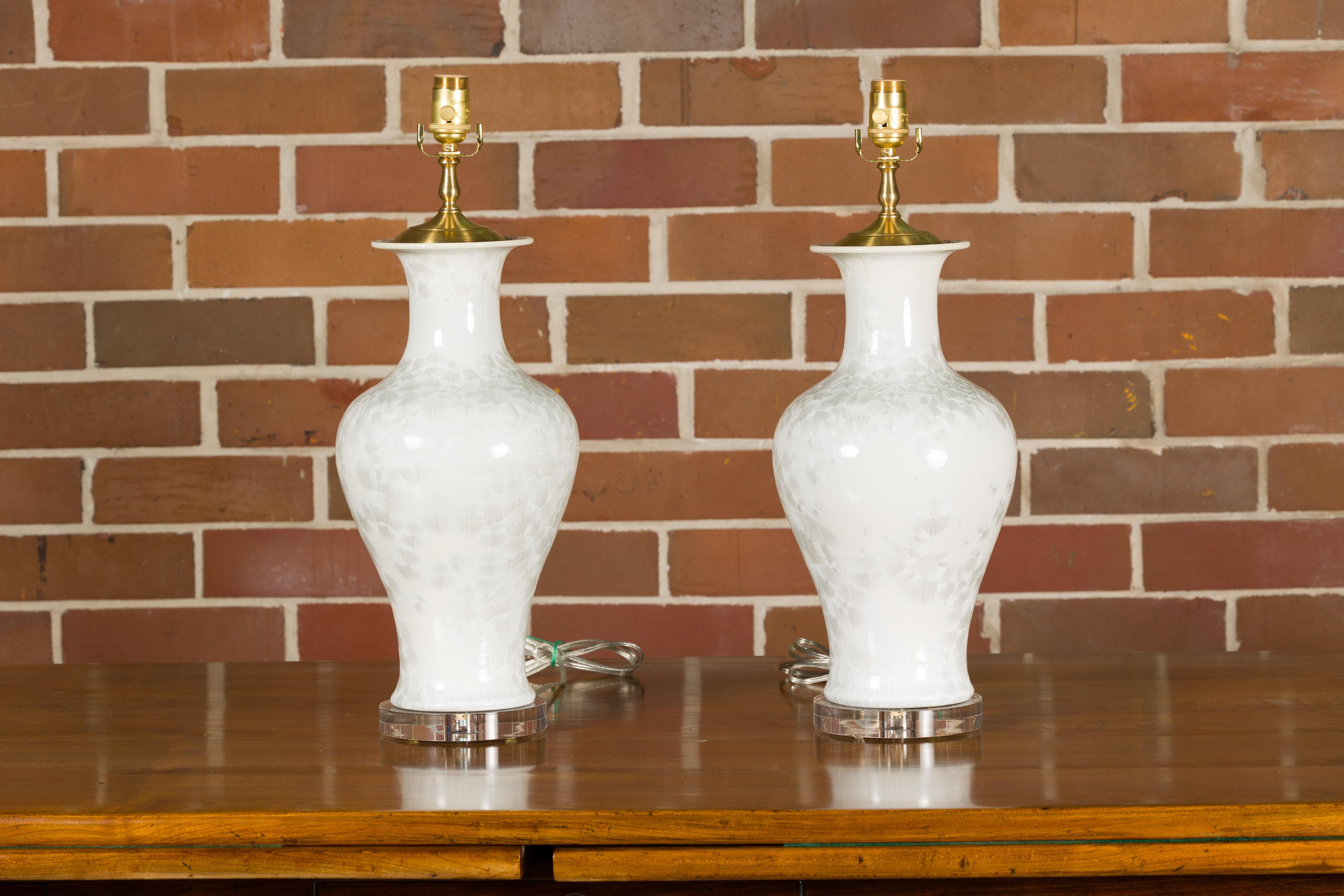 A pair of white porcelain vase shaped table lamps with textured décor and circular lucite bases. Elevate your home's ambiance with this elegant pair of white porcelain vase-shaped table lamps, each boasting a subtle yet exquisite textured décor.