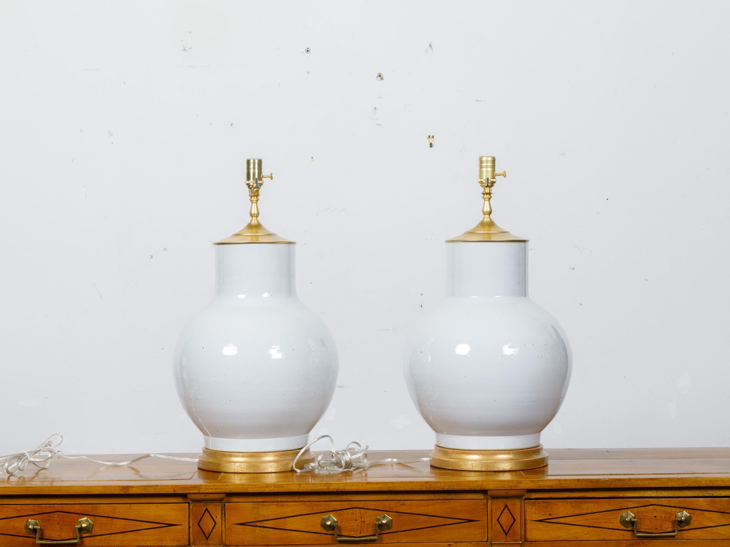A pair of white porcelain vases made into table lamps wired for the USA and mounted on giltwood circular bases. This pair of table lamps, transformed from elegant white porcelain vases, is a splendid fusion of classic aesthetics and modern
