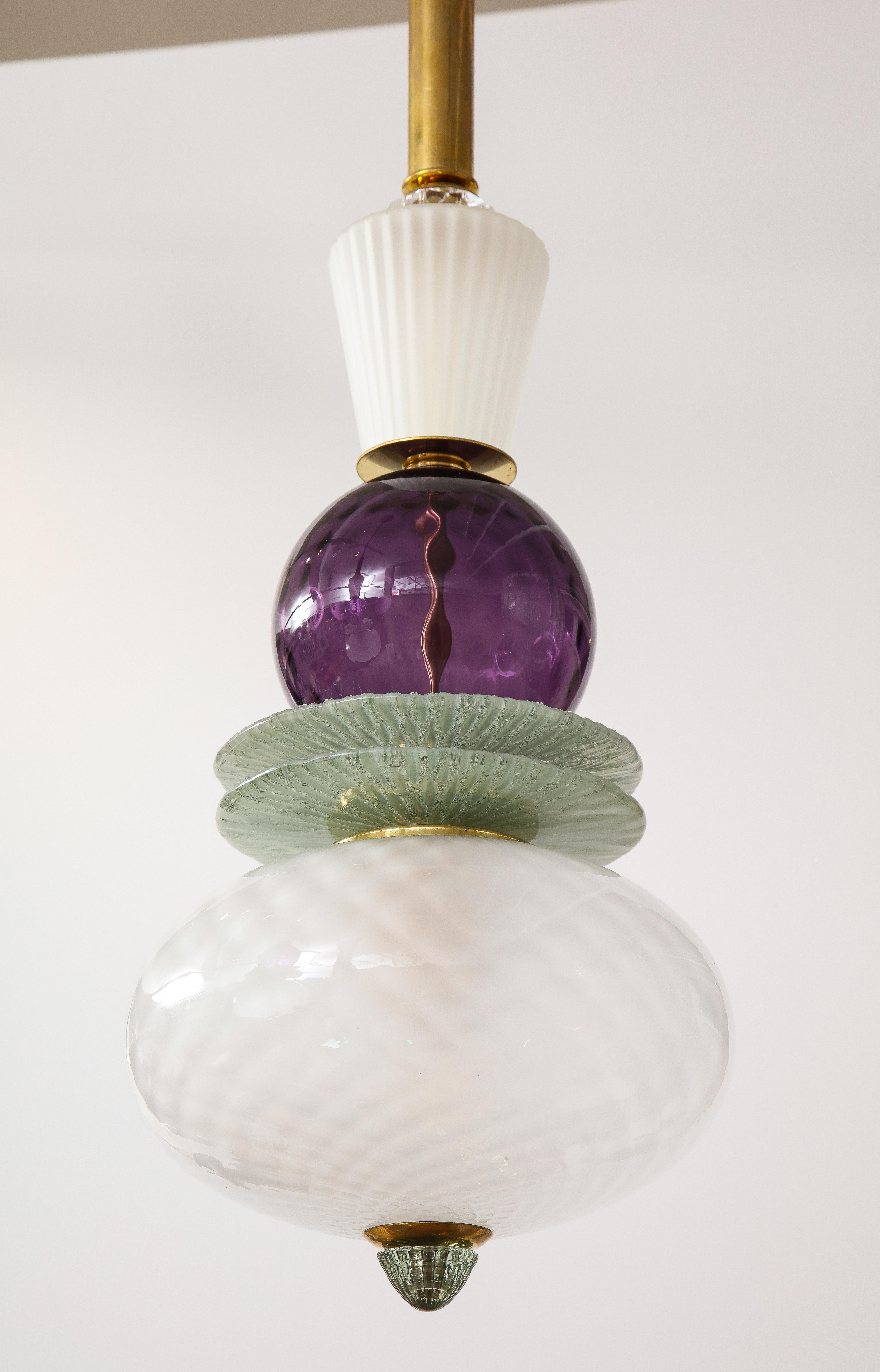 One of a kind pair of Murano glass globes and brass pendants in hues of white, soft green, and purple, handcrafted in Venice, Italy, 2022. A frosted and textured Murano glass globe is stacked with various Murano glass decorative elements, including