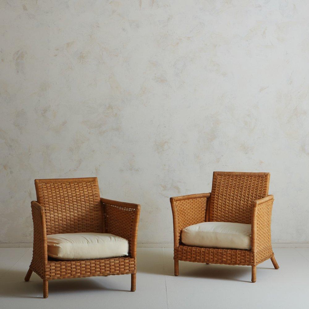 A pair of 1970s French lounge chairs with woven wicker frames. These chairs feature square seat backs with stately arms and angled legs. They retain their original white cushions. Sourced in France, 1970s.