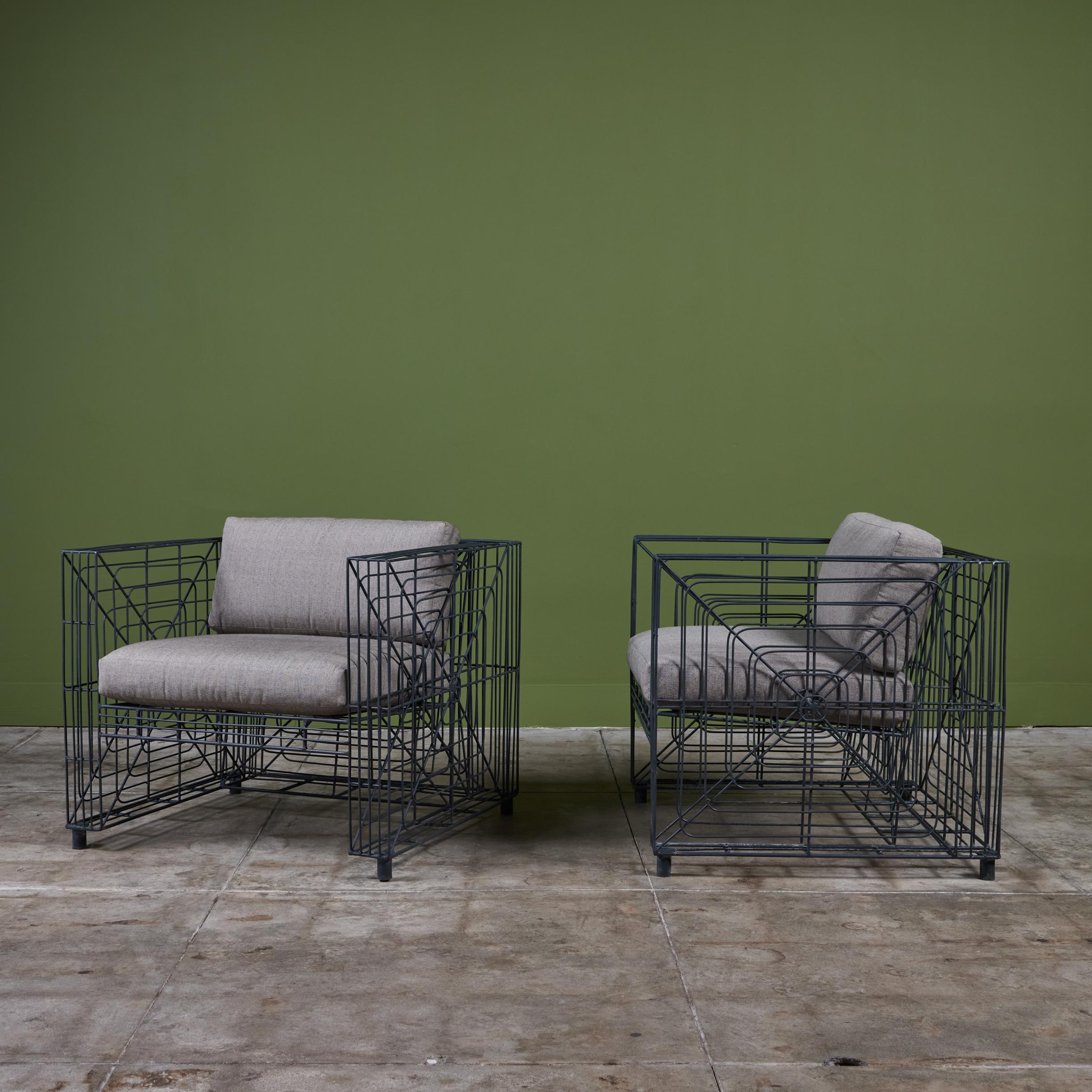Un par de tumbonas de metal para patio. Cada silla tiene forma de cubo con reposabrazos altos y asiento profundo. Cada estructura de alambre muestra un diseño geométrico de tela de araña con un cojín de asiento tapizado y respaldo en gris