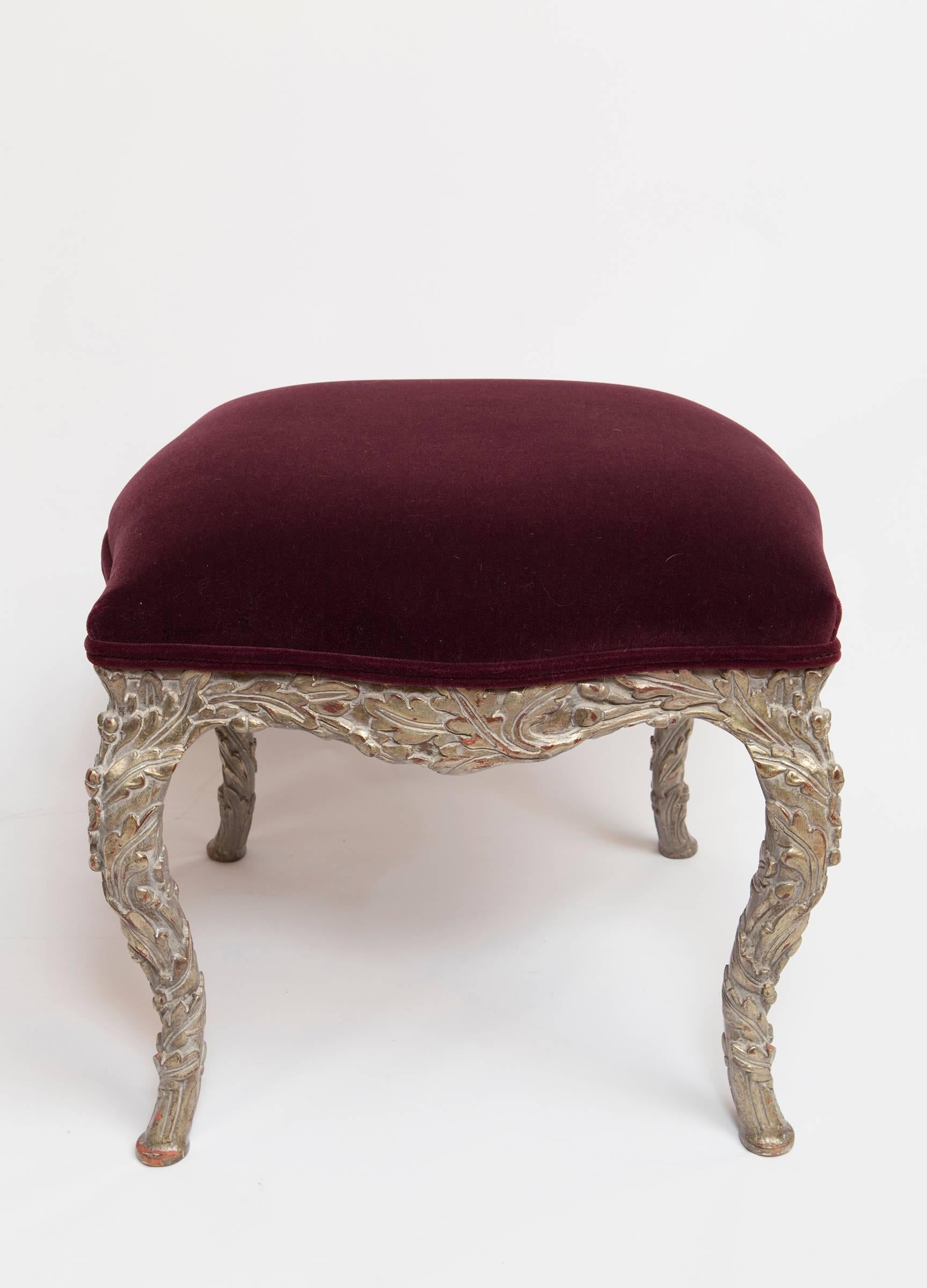 Pair of wood carved stools upholstered in burgundy mohair velvet fabric. The pair of stools are finished in a distressed rubbed silver gilt. The carving is very beautiful in a leaf detail. The size in a generous 21