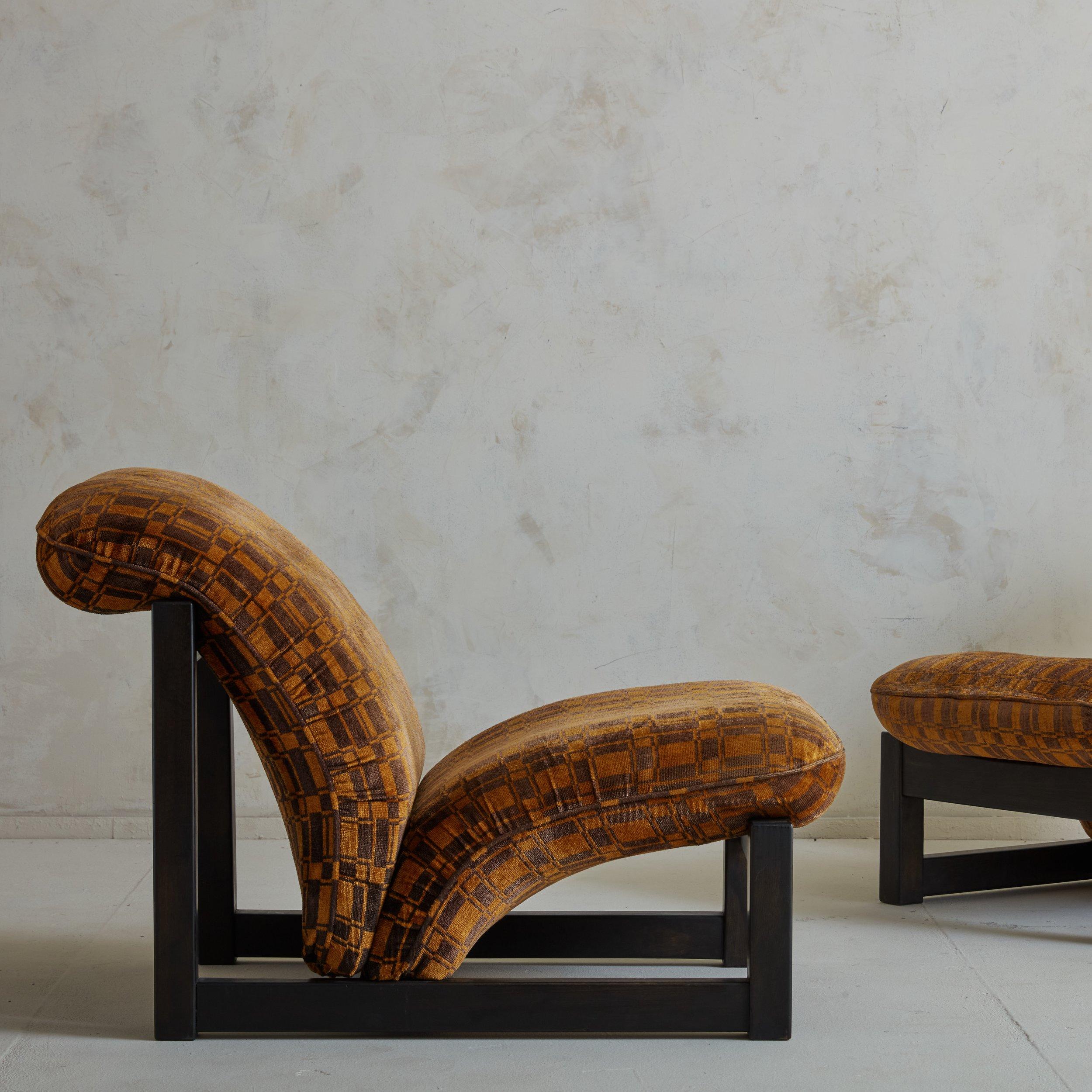 A striking pair of 1970s Italian lounge chairs featuring angular wood frames stained a rich chocolate brown hue. These chairs have dramatically curved seats and seat backs, which retain their original burnt orange and plum checkered velvet