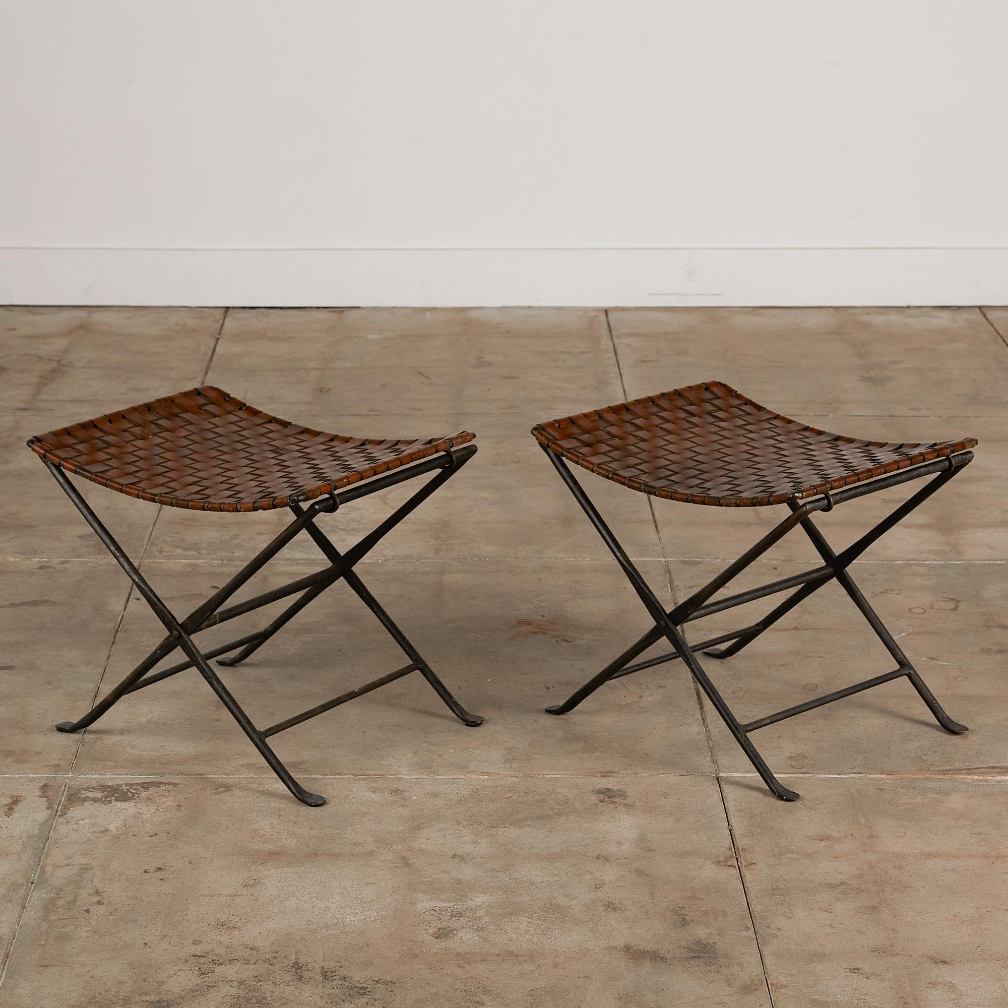 A pair of woven leather stool featuring a folding wrought iron base with an x-frame profile and hammered metal details. The seat is composed of brown leather in a plain weave. The leather straps wrap around the metal frame showing off simplistic but