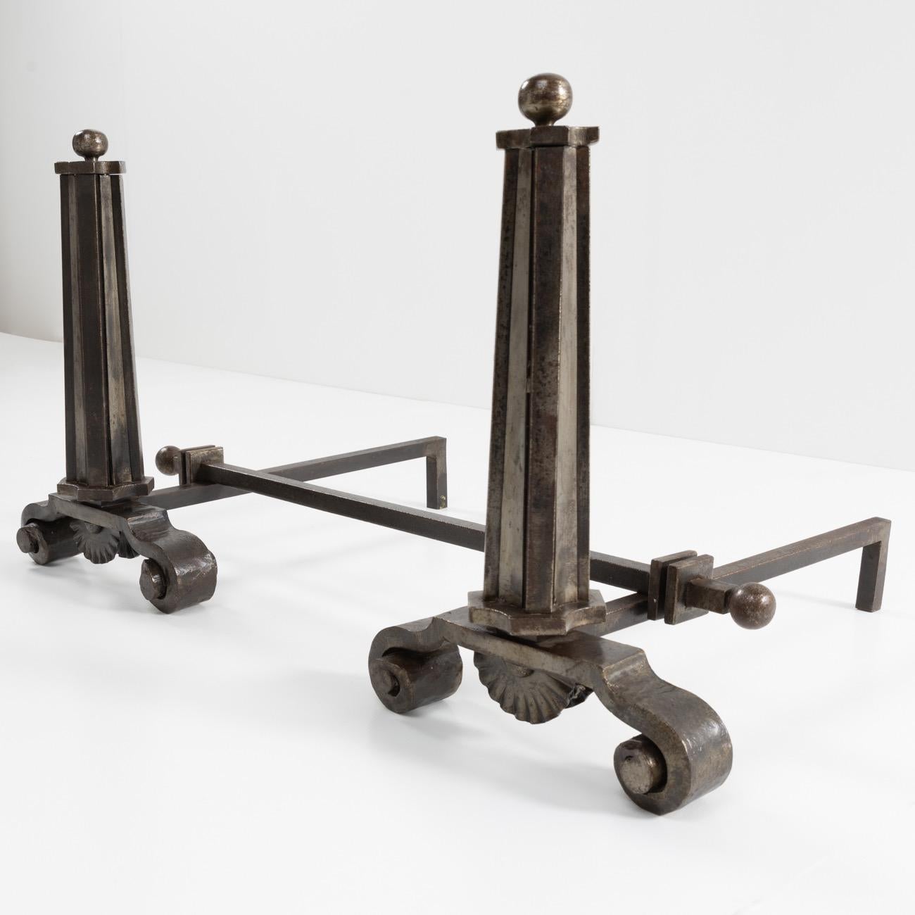 Magnificent pair of Art Deco wrought iron andirons. The frontal column shape surmounted by a decorative ball. The cross bar is mobile. Each andiron signed under the shell 