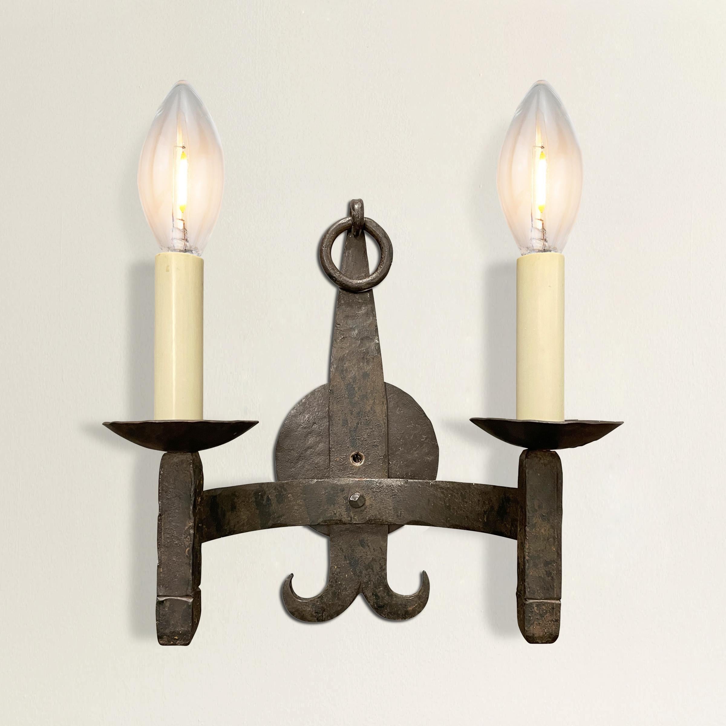 A fabulous pair of mid-20th century American handwrought iron two-arm sconces with square candles held by curved arms, and round back-plates with rings hanging from the tops, and curled bottom ends. Wired for US, and utilizing candelabra size bulbs.