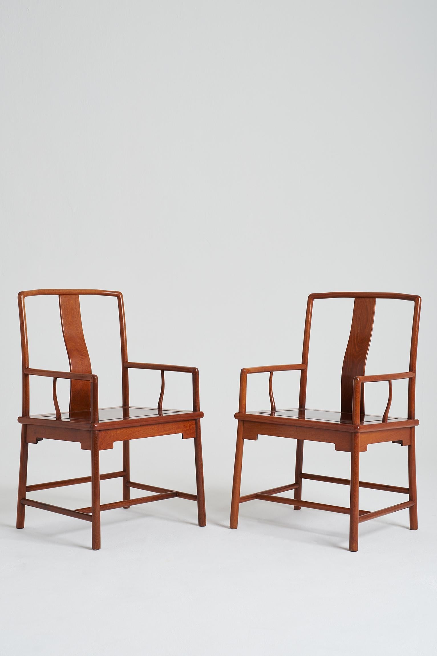 A pair of continuous yokeback armchairs in the Ming style, each chair with an subtly arched yokeback toprail supported by shaped spandrels and curving down to join the gently sloping rear posts continuing through the frame to form the back legs,