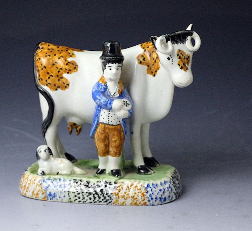 A pair of Pottery Prattware cows modeled standing with a farmer, his wife, dog, and a calf. The cows are decorated with burnt orange patches with black speckles. The female figure is wearing a low shoulder dress and stands with a calf lying at her