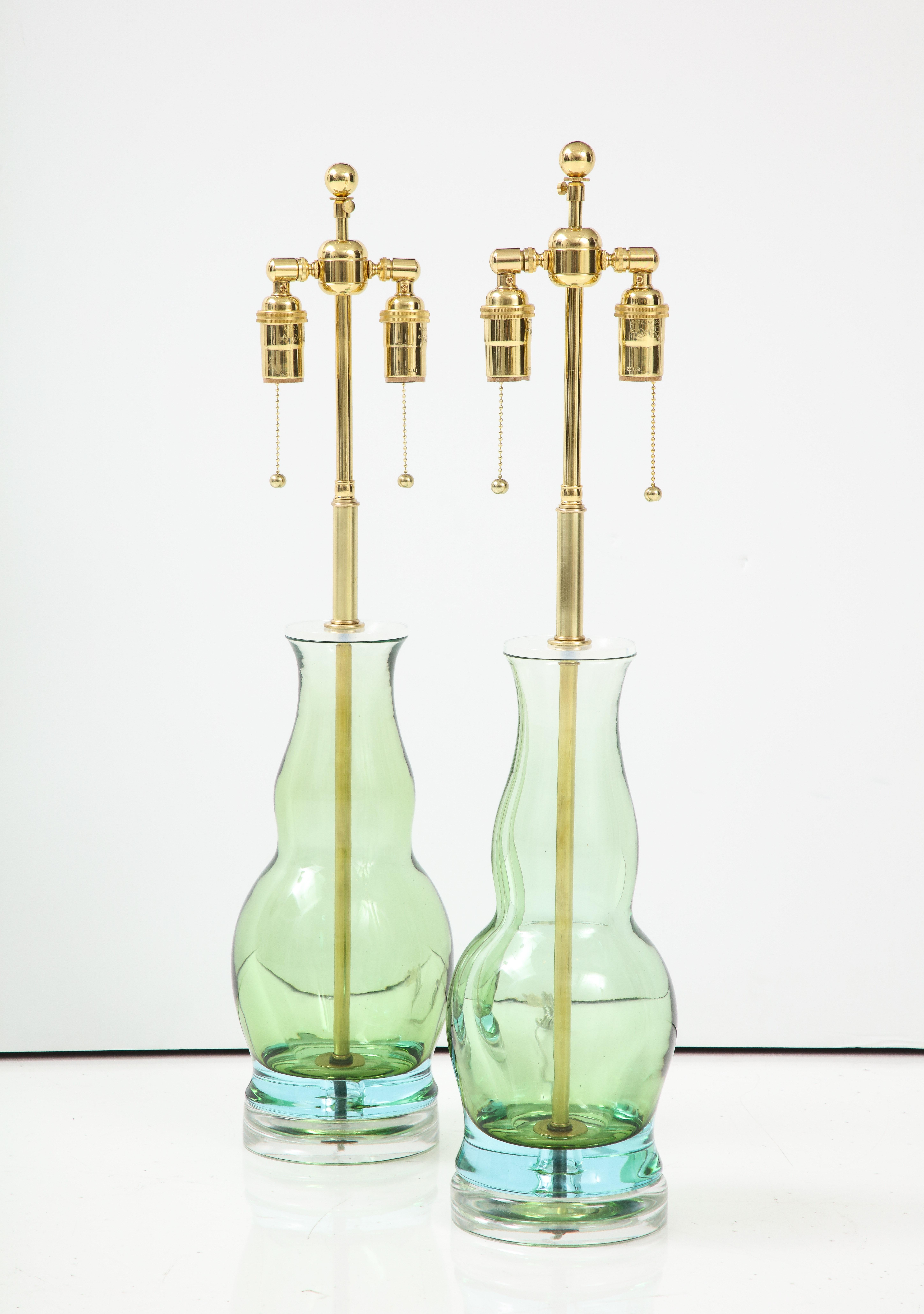 Pair of Bottle form green glass shaped lamps mounted on a thick 
clear glass bases.
They have been Newly rewired with adjustable polished brass double clusters that take standard size light bulbs.
One of the glass forms is slightly larger as they