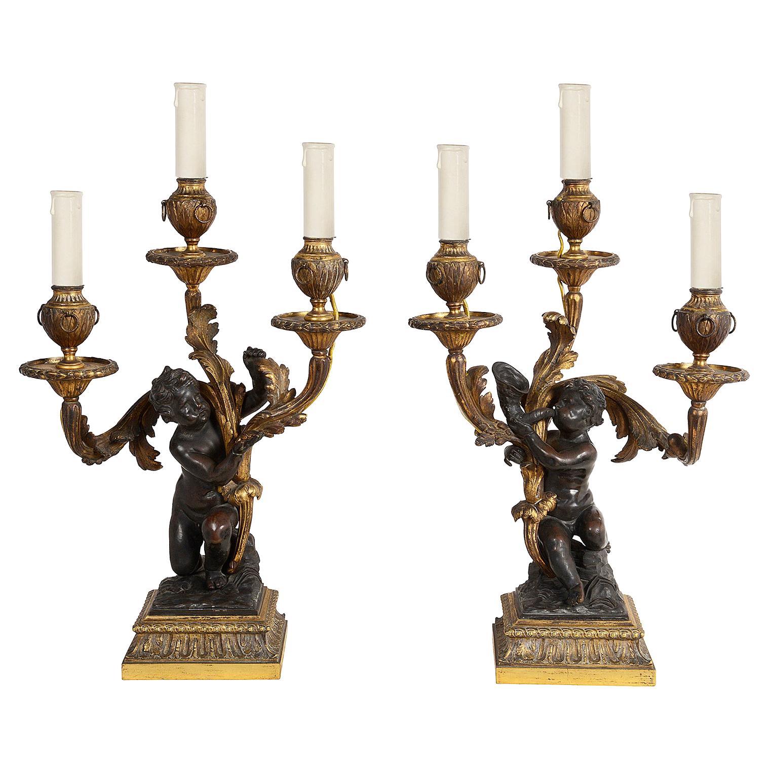 A pair of 18th century style gilded ormolu and patinated bronze three branch candelabra, each with a classical kneeling putti supporting scrolling foliate candelabra with urn like sconces and ring drop decoration, circa 1800.