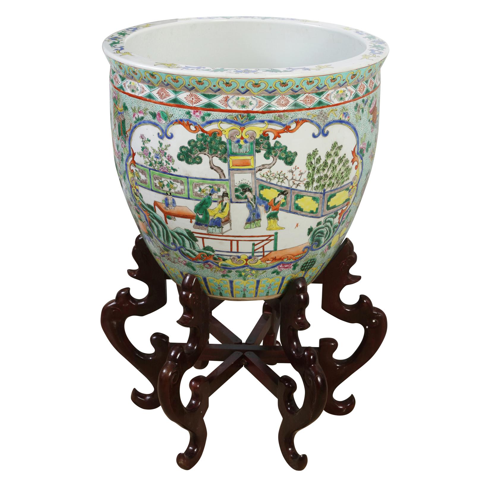 A pair of oversize chinoiserie painted porcelain planters on hexagonal curved wood stands.  The vintage porcelain fish bowl jardineres are decorated with varied shades of greens and blue with two panels of colorful figural and garden scenes. The