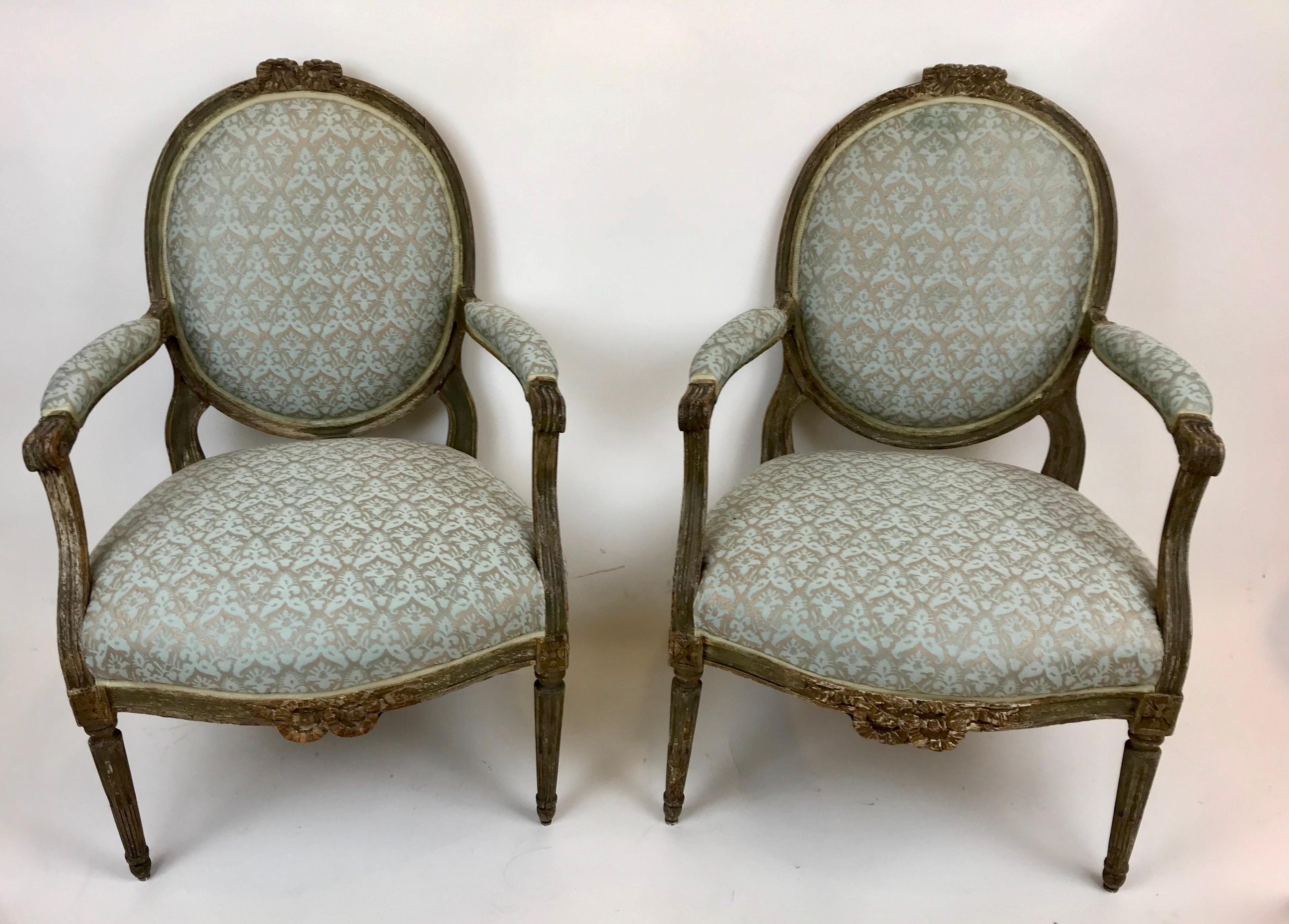 This handsome pair of provincial oval back chairs feature carved bowknots, and stop fluted tapered legs. They are currently covered in floral Fortuny fabric.