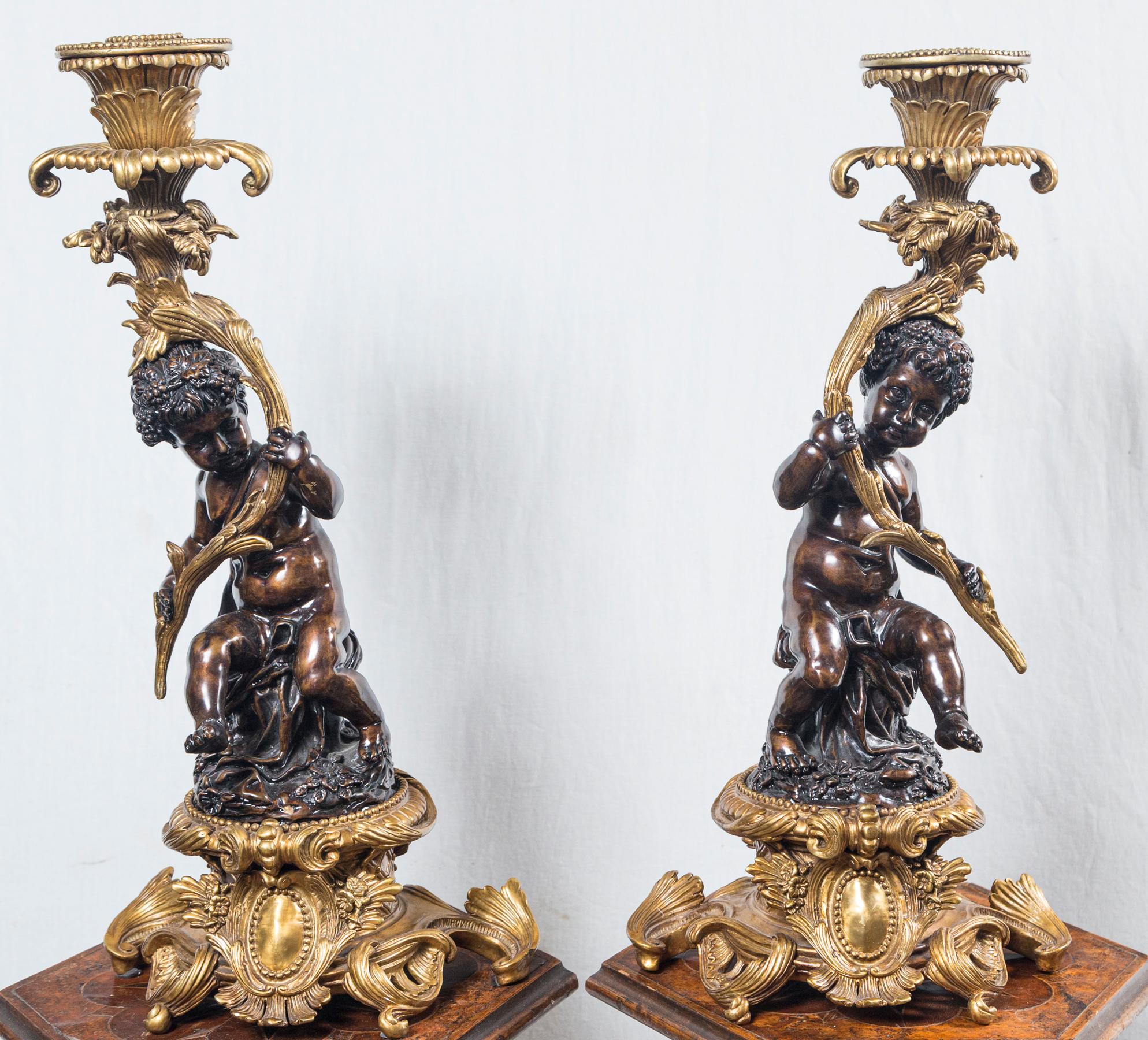 Rococo style gilt bronze base, each putto sits on a patinated rock form base. They hold a tall branch that supports the candle cup and baubeche at the top. Grape clusters adorn their hair.
The putti are very much, stylistically, in the manner of