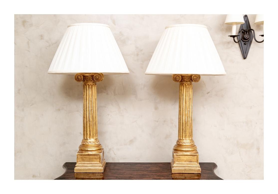 Pair of large- scale gilt ionic column form twin-light table lamps on square pedestal bases. Each with carved decorative bands of details at the tops and around the round bases. With lined pleated silk shades. The lamps with dual pull chain