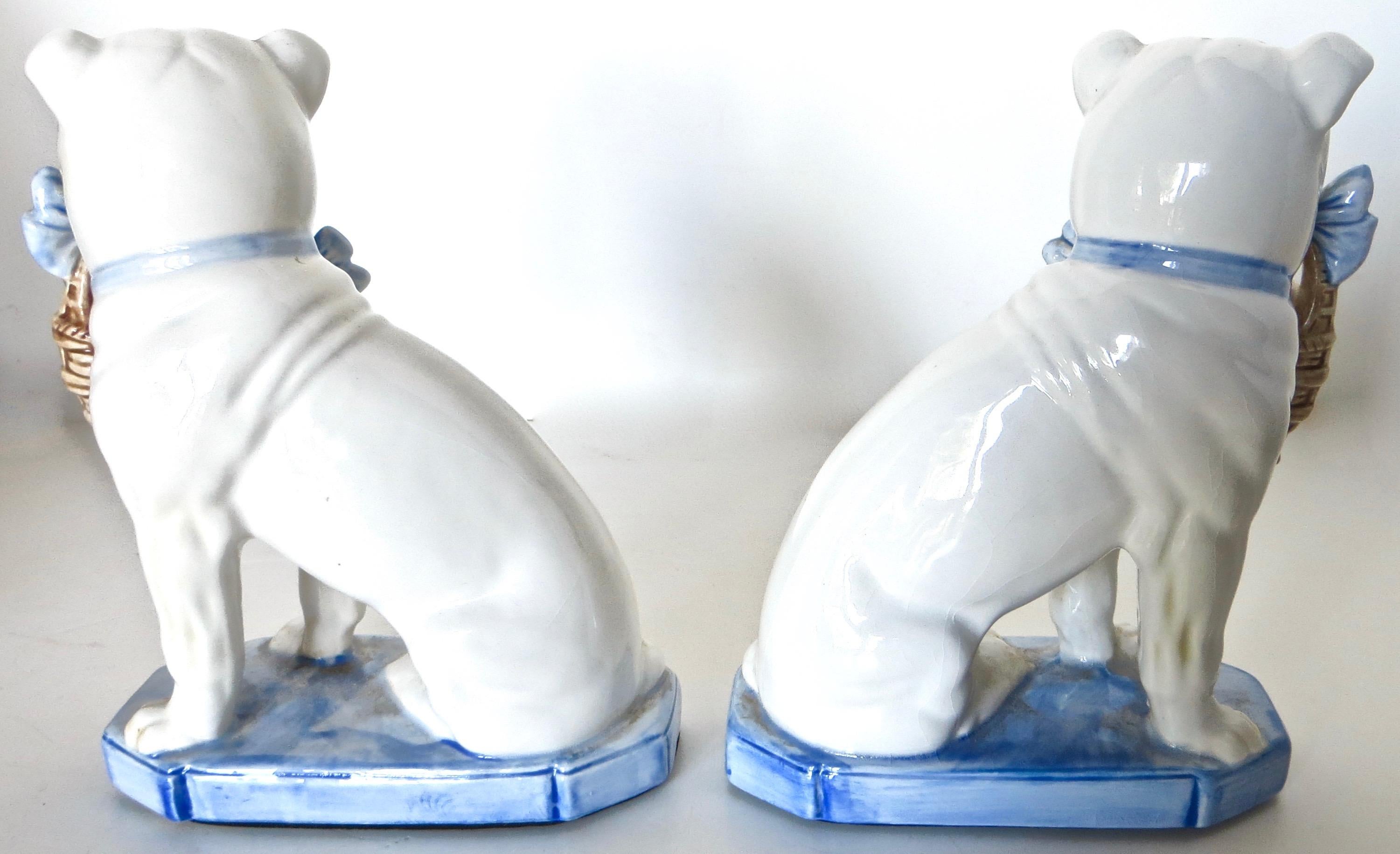 Charming pair of porcelain white seated Bulldogs holding a pair of puppies each from their mouth in a straw basket with a blue bow around their necks; mounted on a blue base. Condition is like new with no cracks or chipping. American manufacturer is