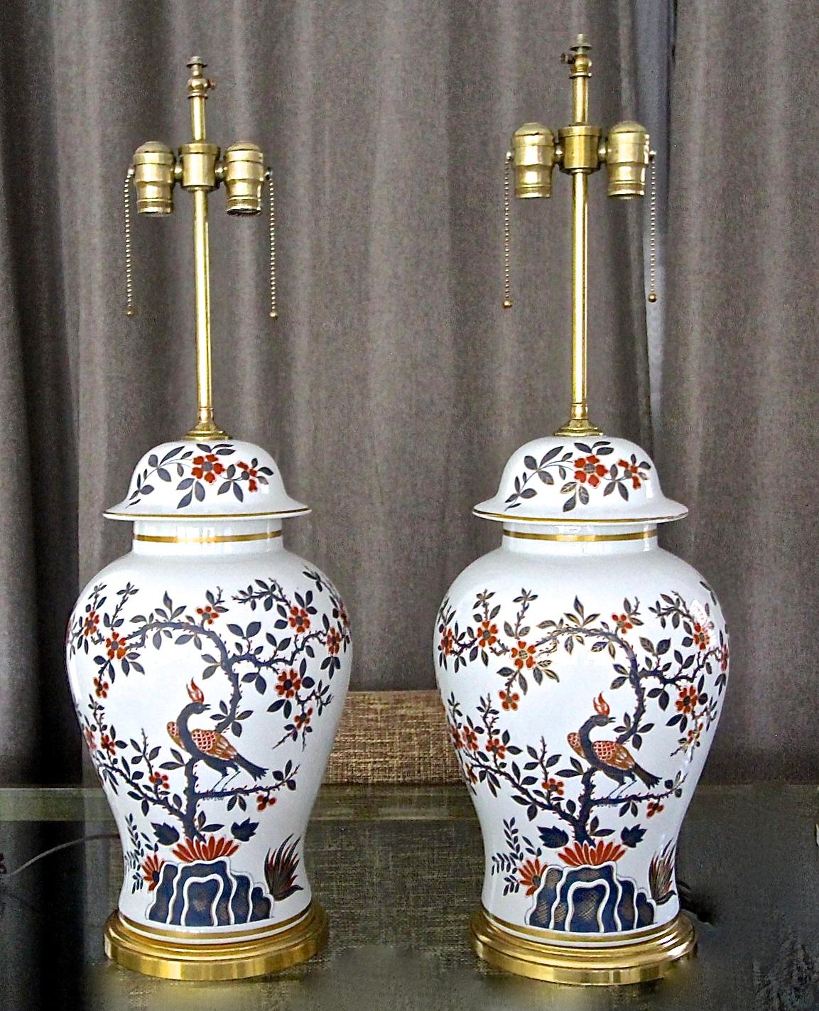 Pair of French porcelain lidded vase Chinoiserie style table lamps resting brass bases. Nicely detailed with peacock bird and floral motif. Hand painted in red and blue with gold highlight trim. Each with double cluster brass pull chain sockets and