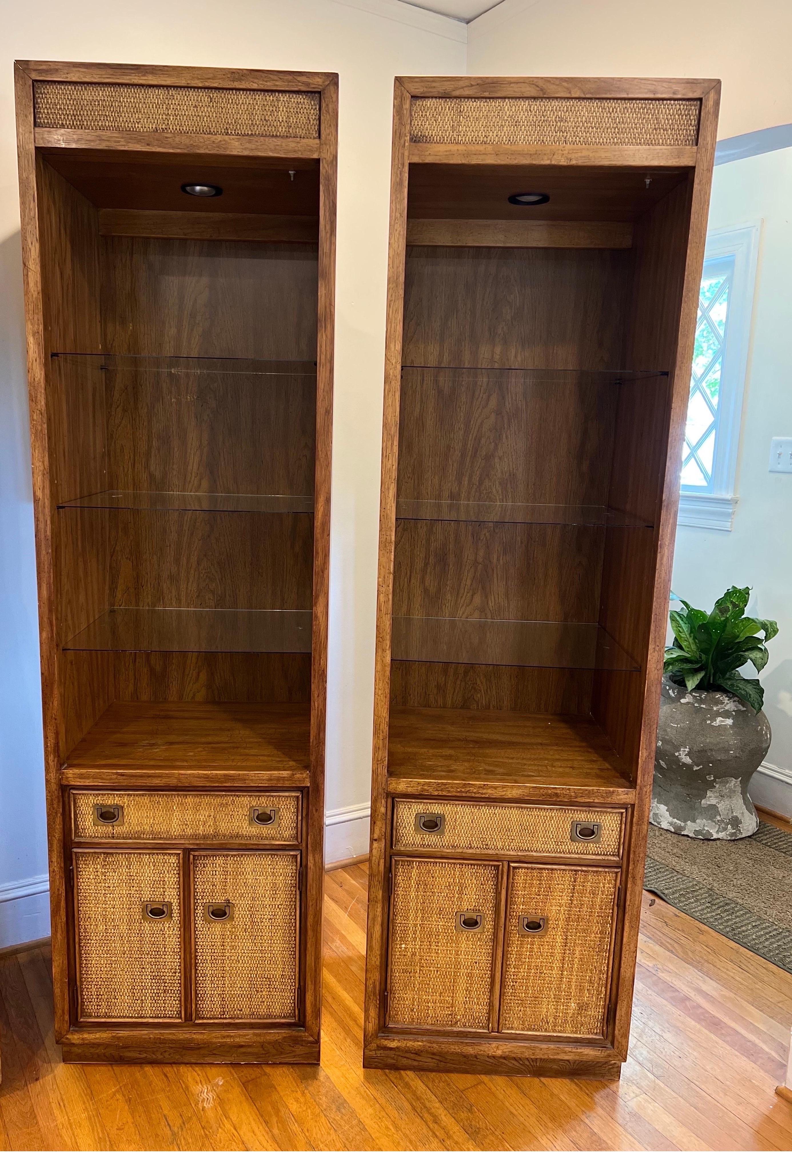 Rare pair of Mid-Century Modern Campaign style case pieces/display shelving.
Walnut frame with rattan details. 
Each has a single drawer and doors that open to an area with a shelf. 
Glass shelving. Wired for lighting, but needs repair. 

See