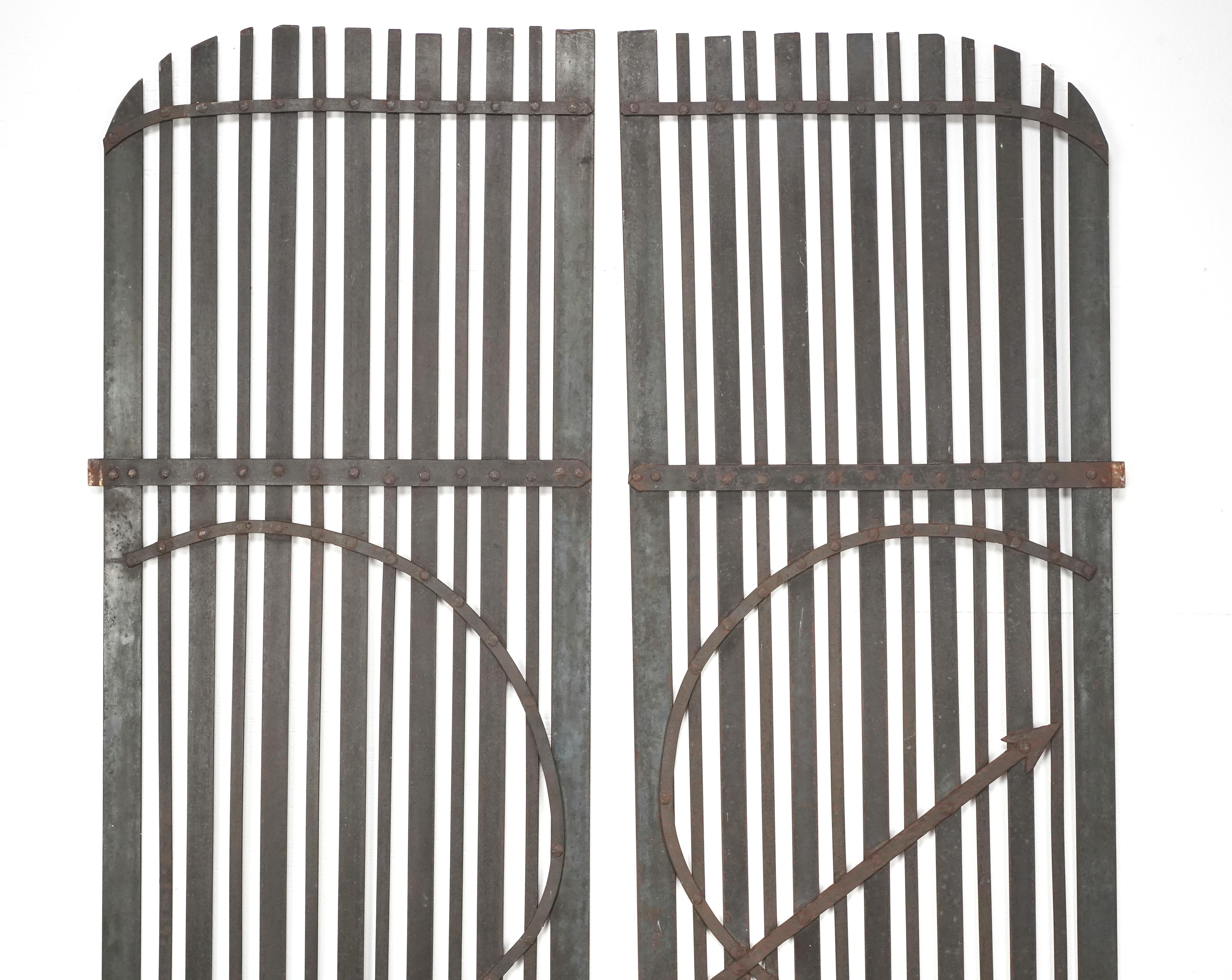 Reclaimed arched wrought iron gates featuring an interlocking circular design bisected by a stylized arrow and horizontal supports. Good condition with surface wear and rust from prior use. Priced as a pair. One pair available. Please note, this