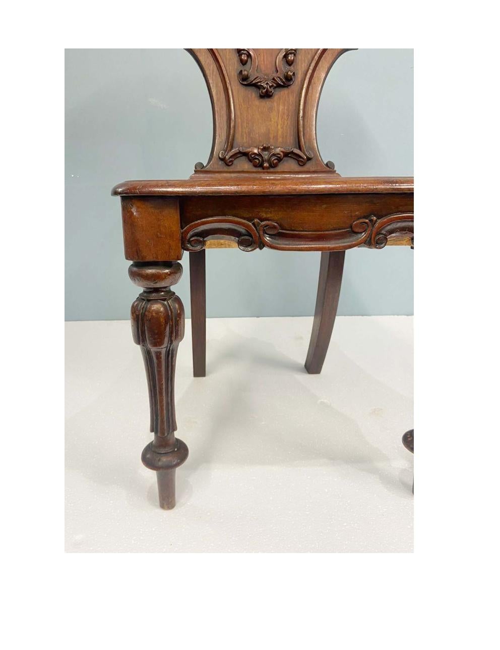You are viewing a gorgeous pair of period Regency hall chairs with Gillows motifs
Each chair features a solid, shield-shaped backrest adorned with high-relief carving, including acanthus leaf motifs, scrollwork, and a central heraldic