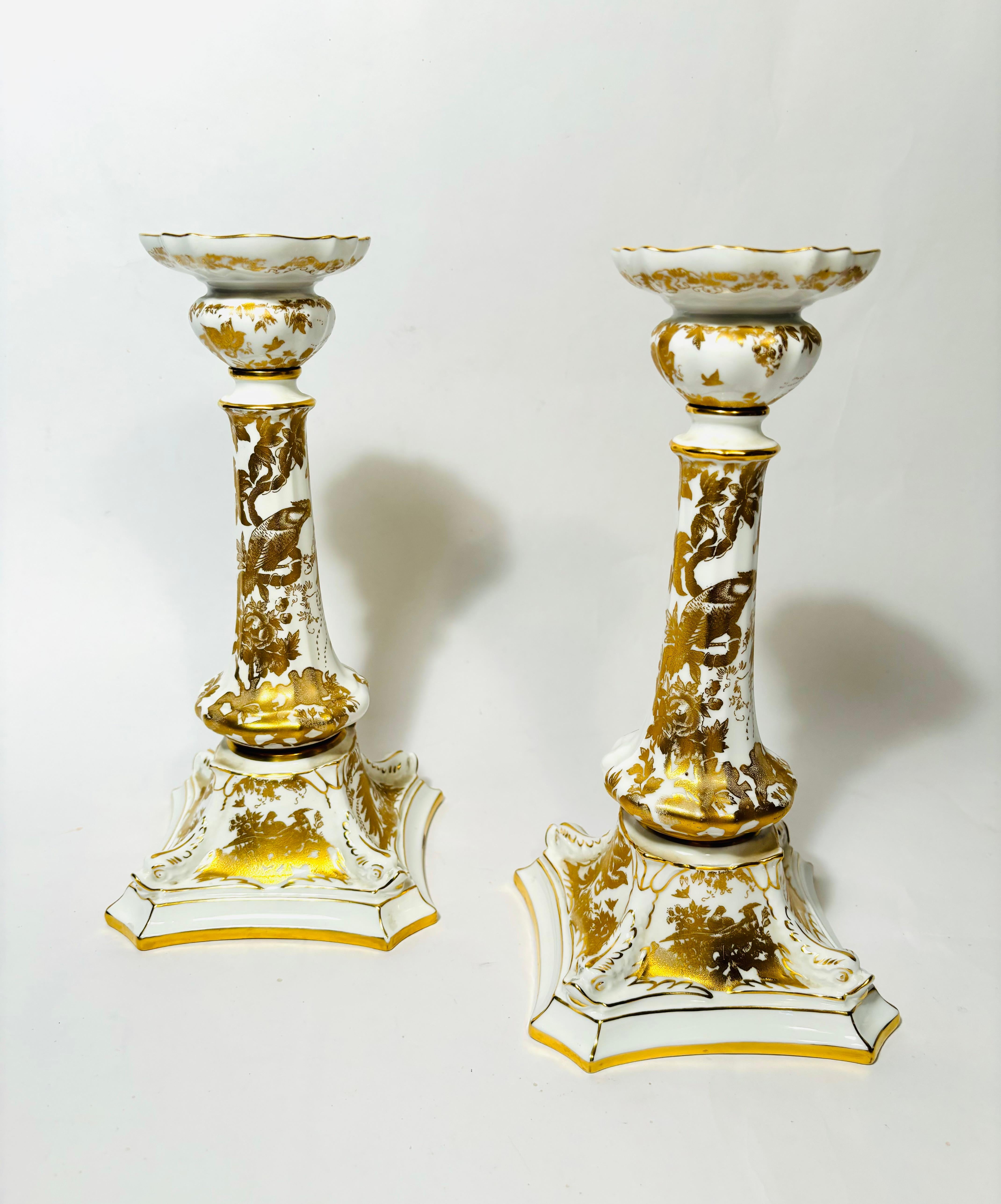 An elegant pair of 24 karat gold and white fine porcelain candlesticks by the re known English firm of Royal Crown Derby. This elaborate design of adorned birds and peacocks is applied throughout the pieces. The Aves pattern was adapted from an