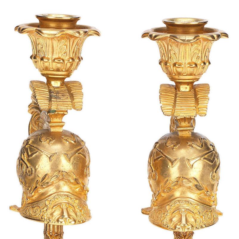 Our pair of ormolu bronze candlesticks feature acanthus leaf nozzles emanating from Roman style military helmets with plumage, with reeded acanthus stems and laurel swags with shields, and are raised on stepped square bases with wreath motifs.