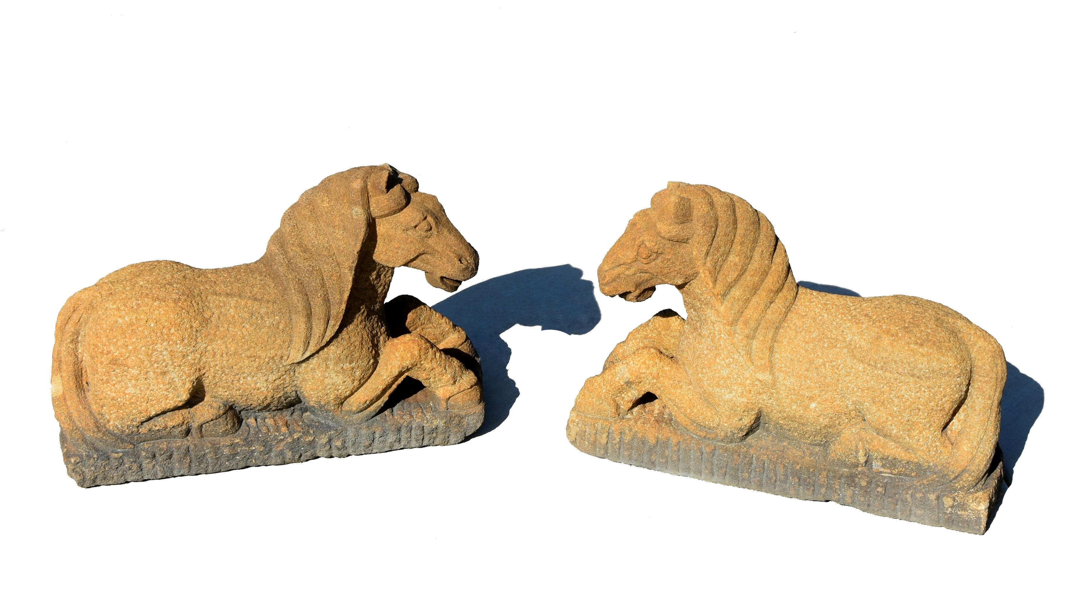 A pair of hand carved, large solid stone horse statues. The recumbent horses are crafted with two legs tucked under the body and tail flicked to one side. The well incised manes drapes around the heads behind the forward-pricked ears. The muscular