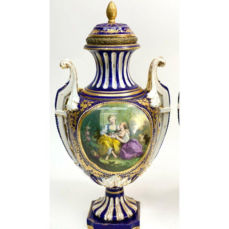 Pair sevres France porcelain twin handled covered urns, 19th century.

A cobalt blue ground with gilt leaf decorations throughout. The central area of the vases depict courting scenes to one area and floral bouquets to the other. White enamel