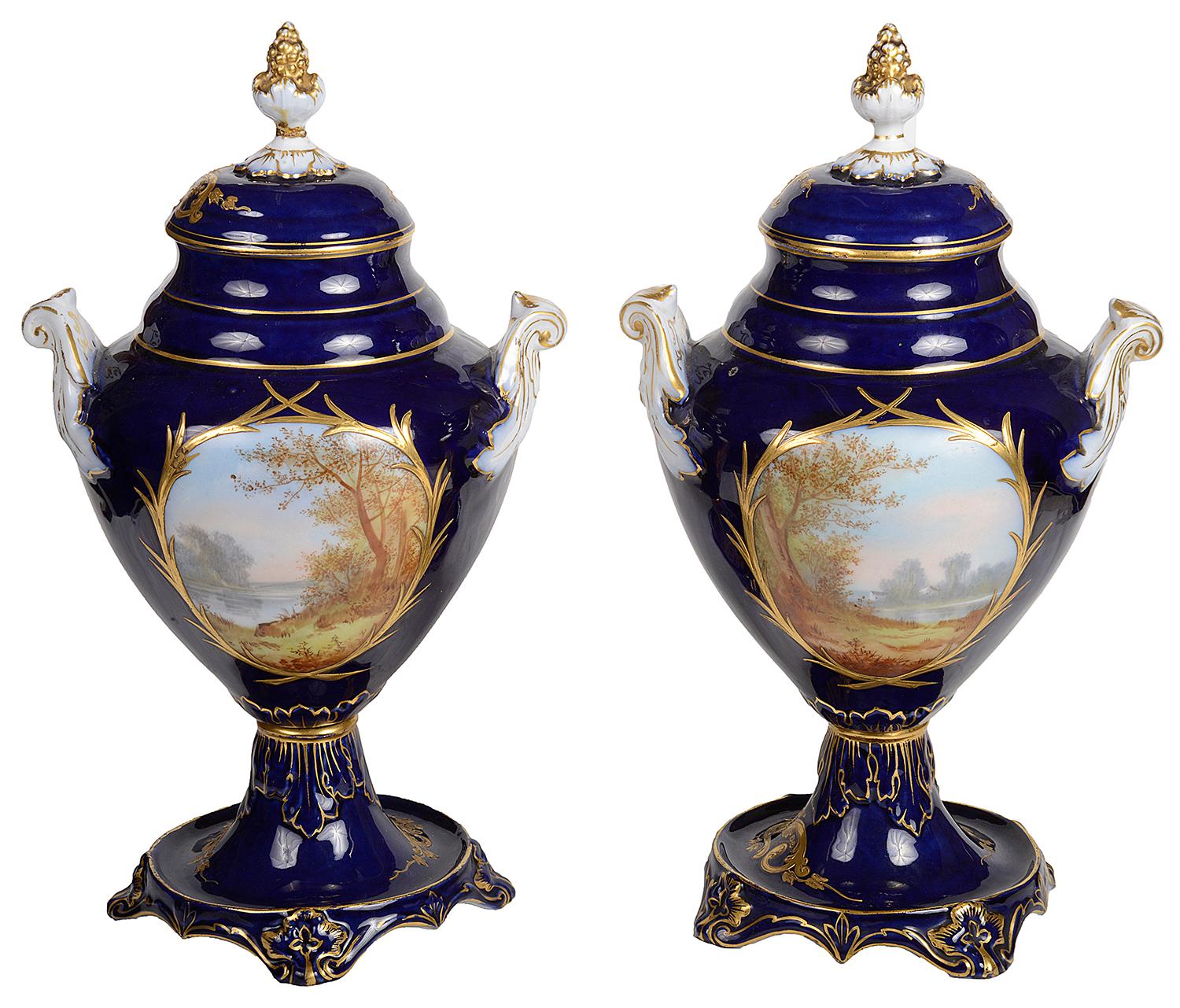 A pair of French Sevres style porcelain lidded vases, each in a cobalt blue ground, berry and leaf finials, classical scrolling gilded decoration, inset painted panels depicting young lovers, scrolling handles to either side. Measures: 38cm/ 15