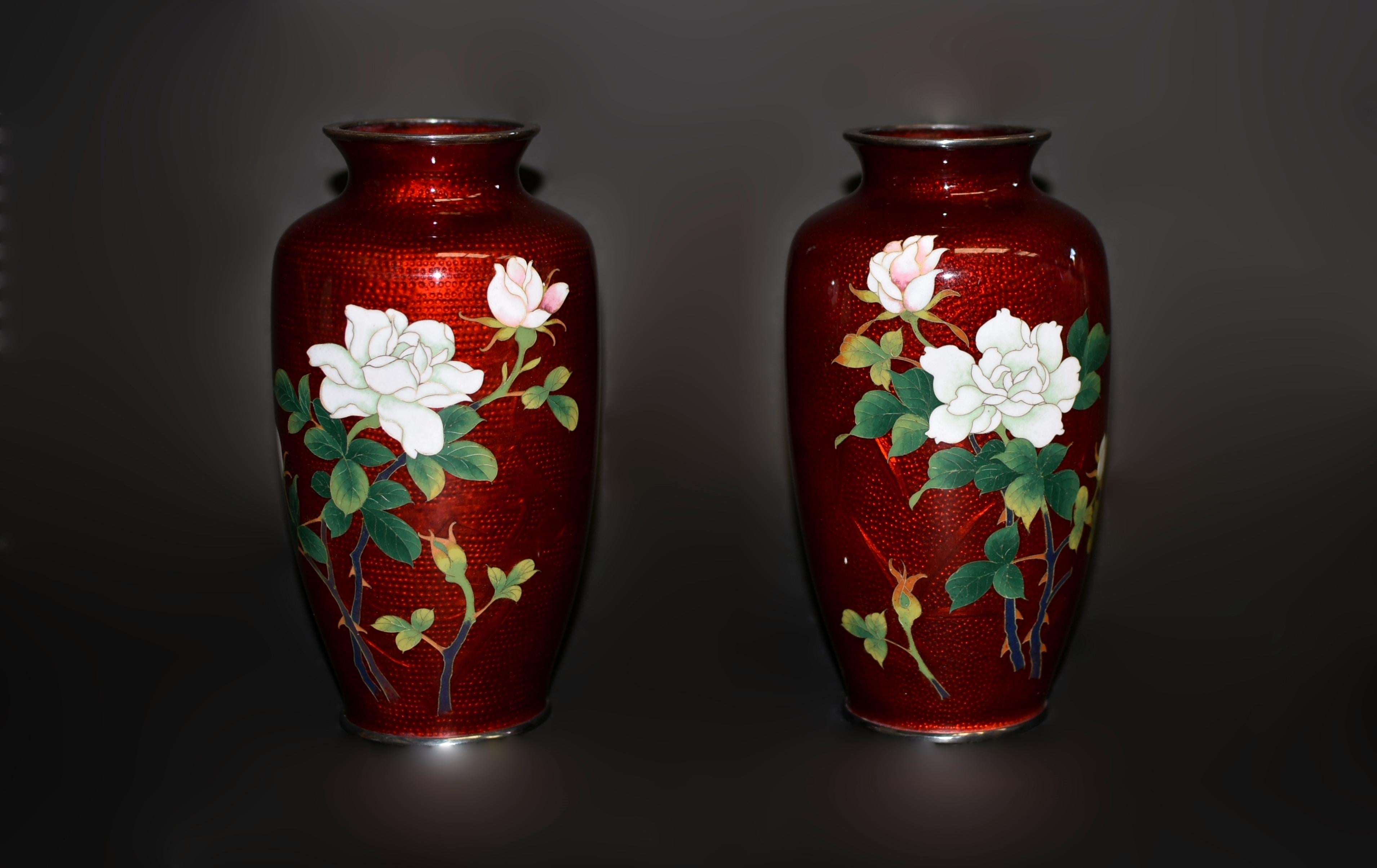 Un par de hermosos jarrones rojos de cloisonné Sato ginbari. En cada cuerpo, dos ramas de rosas blancas en flor y en capullo con follaje verde, trabajadas con minucioso detalle, sobre un impresionante fondo rojo metálico que representa bambú, pino,