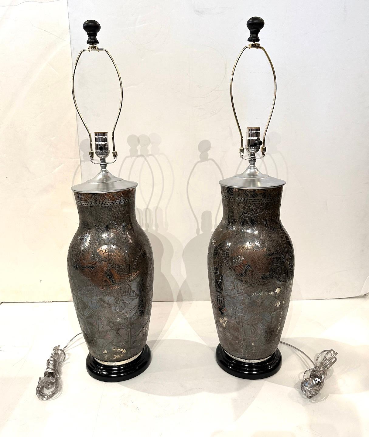 A beautiful pair of silver over copper urns with intricate etched designs of a center bird and floral details later made into lamps.  Single bulb, painted wood base.  Lamps are 32.5