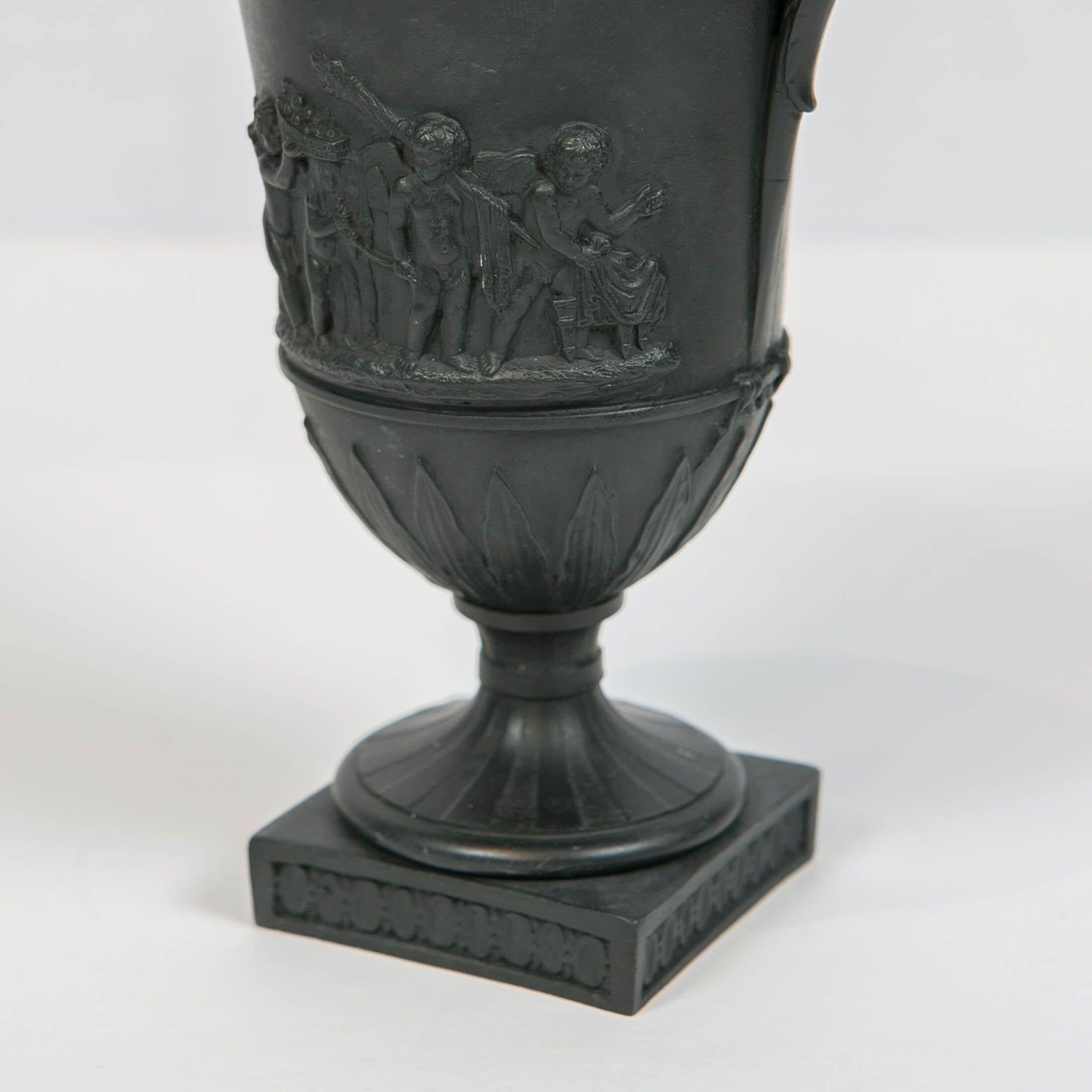 We are pleased to offer this pair of Wedgwood black basalt double handled vases decorated with neoclassical figures. Made in England circa 1800, these covered vases are molded in an ancient Greco-Roman style. Decorated at the top with machine turned