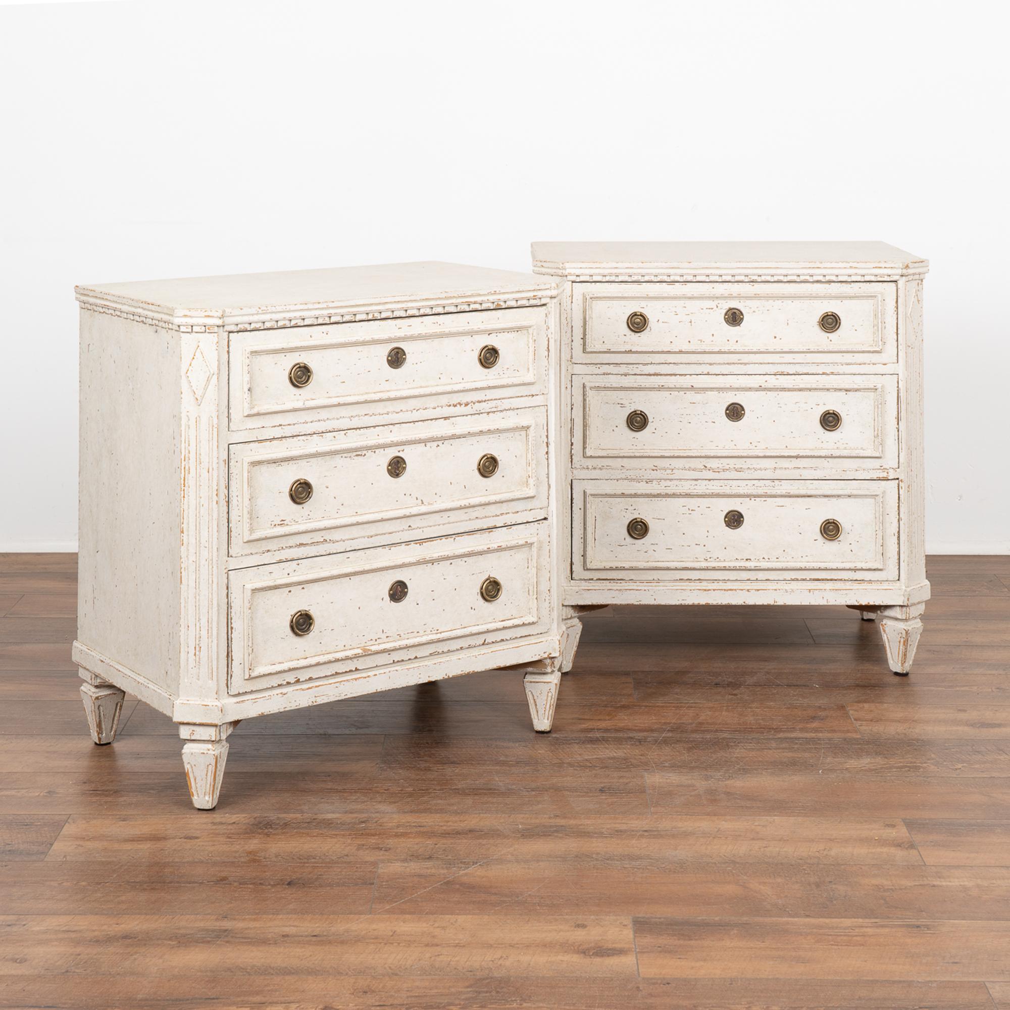 A pair of small Gustavian pine chest of drawers painted in shades of  white, fitting their Swedish origin.
Canted fluted side posts with upper carved diamond medallion, dentil molding, raised carved panels along the three drawers, all setting on
