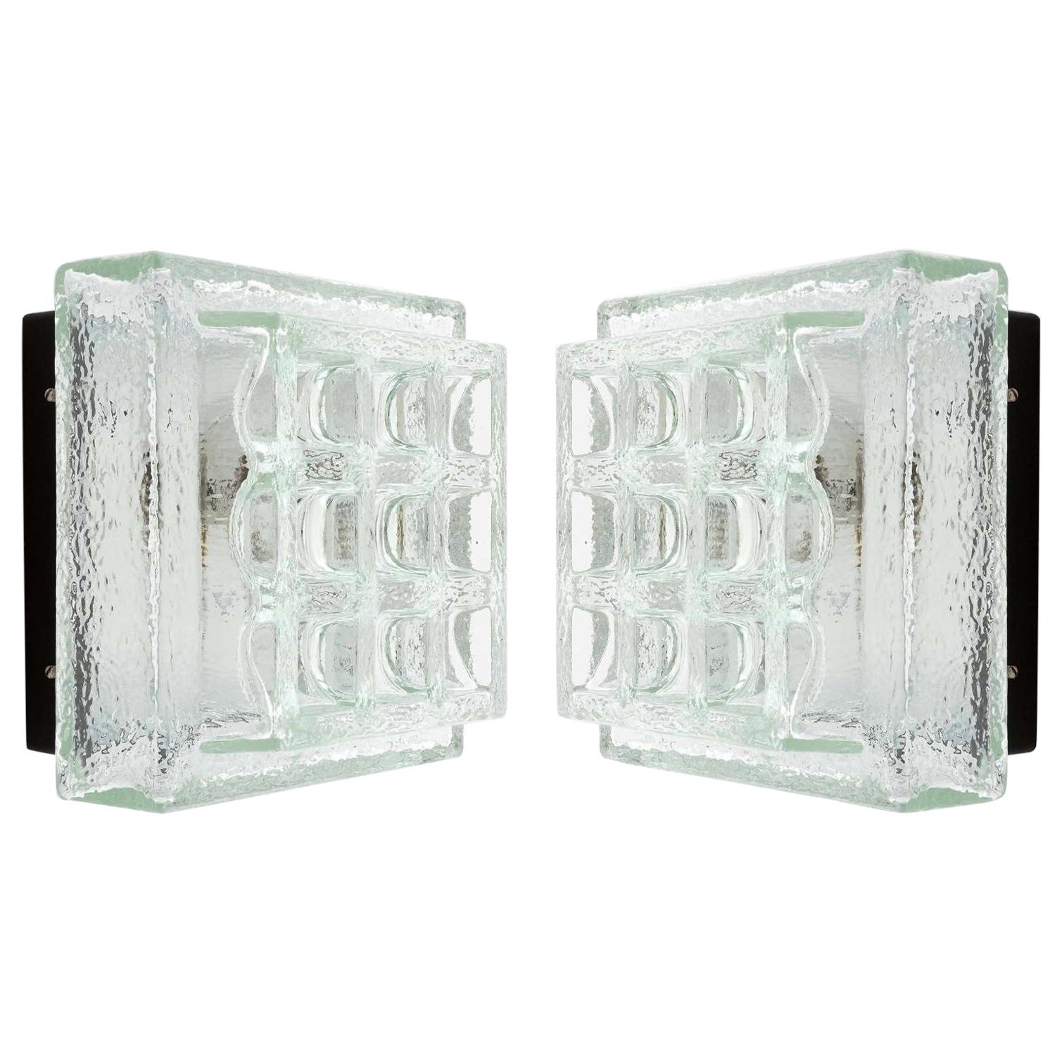 A pair of square modernist flush mount light fixtures or sconces by Glashütte Limburg, manufactured in midcentury, circa 1970 (late 1960s or early 1970s). A black lacquered metal backplate holds a thick geometric textured ice glass. 
The fixtures