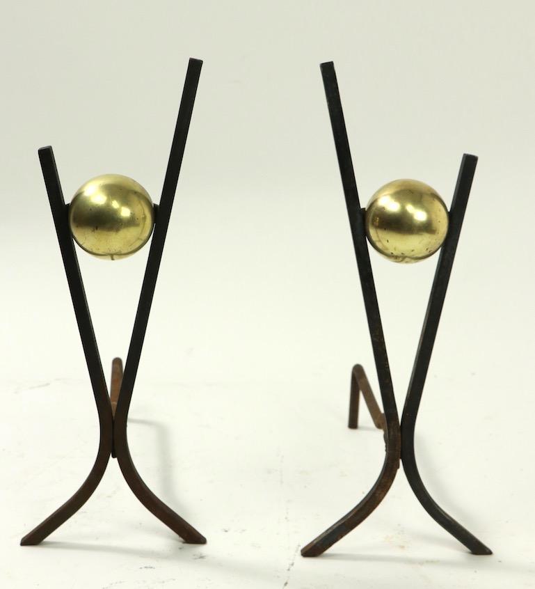 Pair stylish Mid-Century Modern andirons, having squared wrought iron frames which support a dimensional brass ball. Clean, original and ready to use condition, showing only light cosmetic wear, normal and consistent with age.