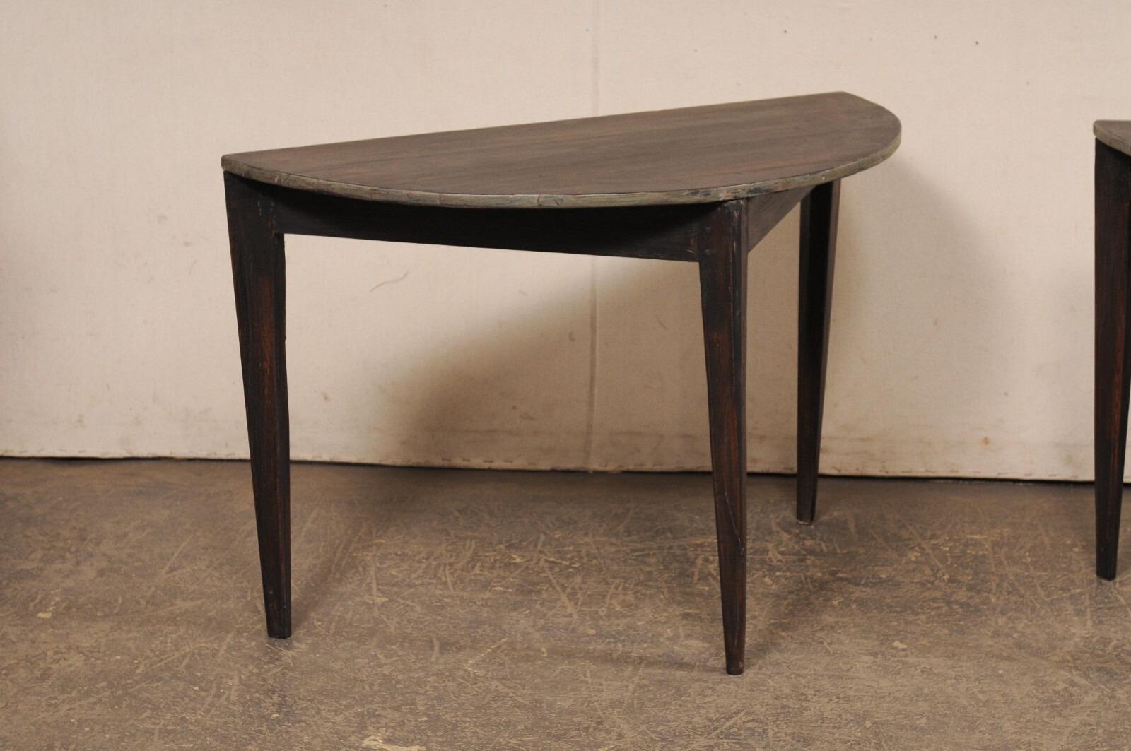A Swedish pair of wooden demi-lune tables from the early 20th century. These antique tables from Sweden each features half moon shaped tops, over triangular aprons, and are raised on three squared, tapered legs. The design is simple and clean. The