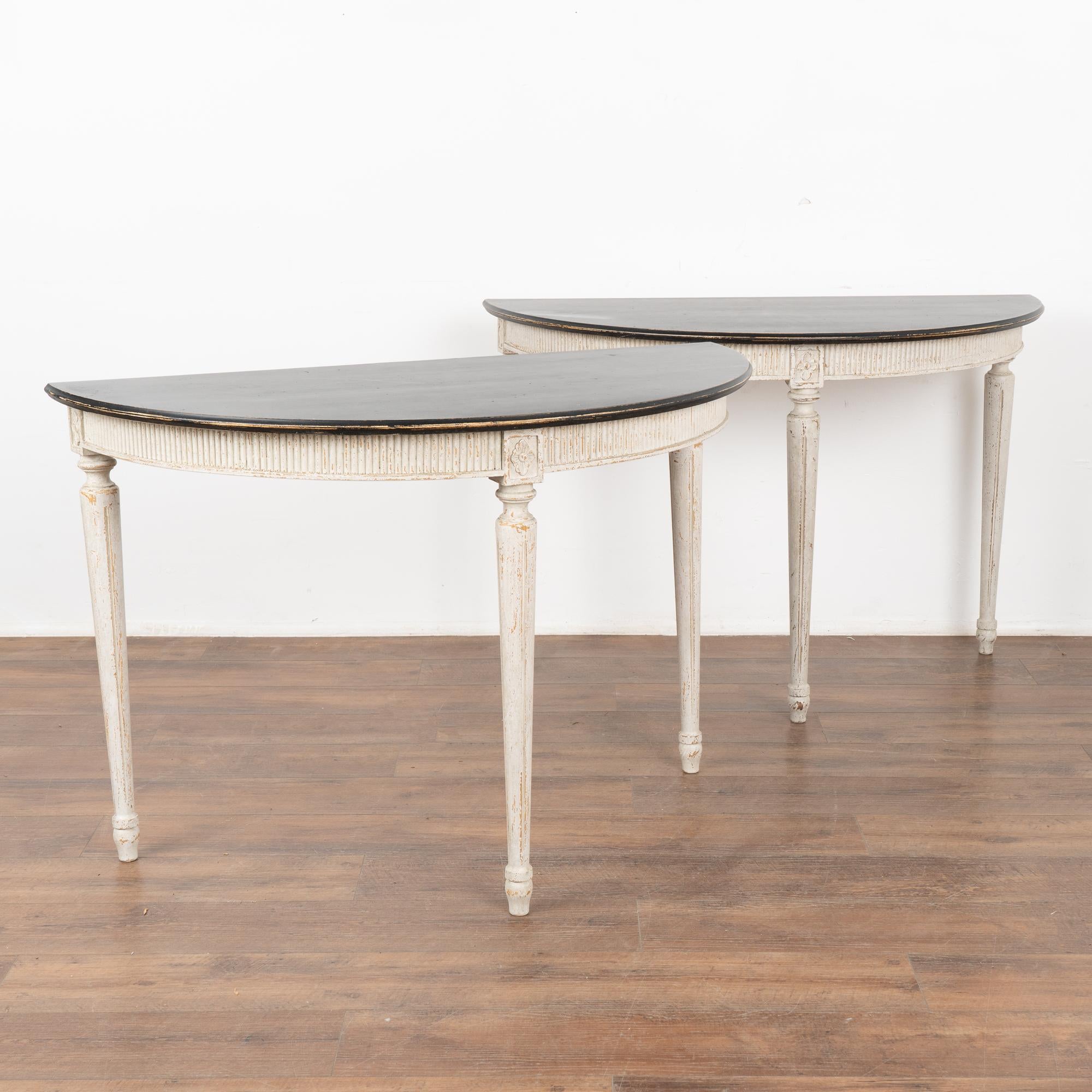 Pair, Gustavian demi-lune pine side tables or consoles with gently tapered fluted legs and decorative fluted carving along skirt.
Newer professionally applied layered antique white painted finish with gray undertones and contrasting black top,