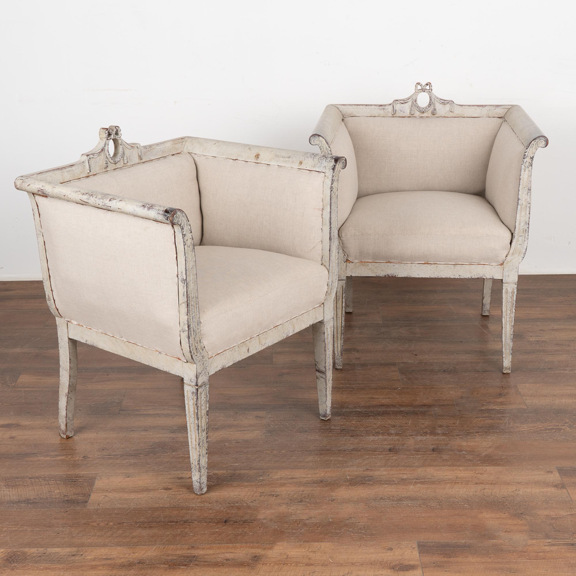This pair of Swedish arm chairs are stunning with their square shape and gently curved arms. A traditional wreath with ribbon accents the back while the fluted detail carving follows the arms down along the tapered legs. 
The original white painted