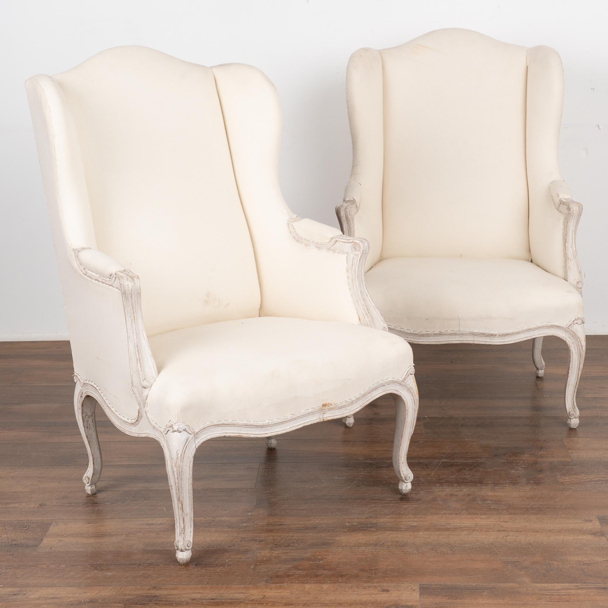 Graceful with a romantic air, this pair of Gustavian style wingback arm chairs still maintain their charming original white paint, lightly scraped revealing a warm patina of natural wood below. 
Simple linen upholstery on back, seat and arms has